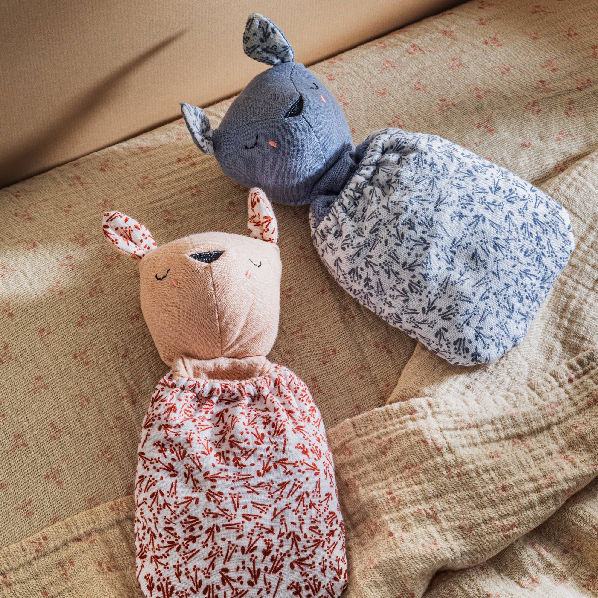 Two soft toys, one pink and one gray with floral patterns, on a textured fabric surface.