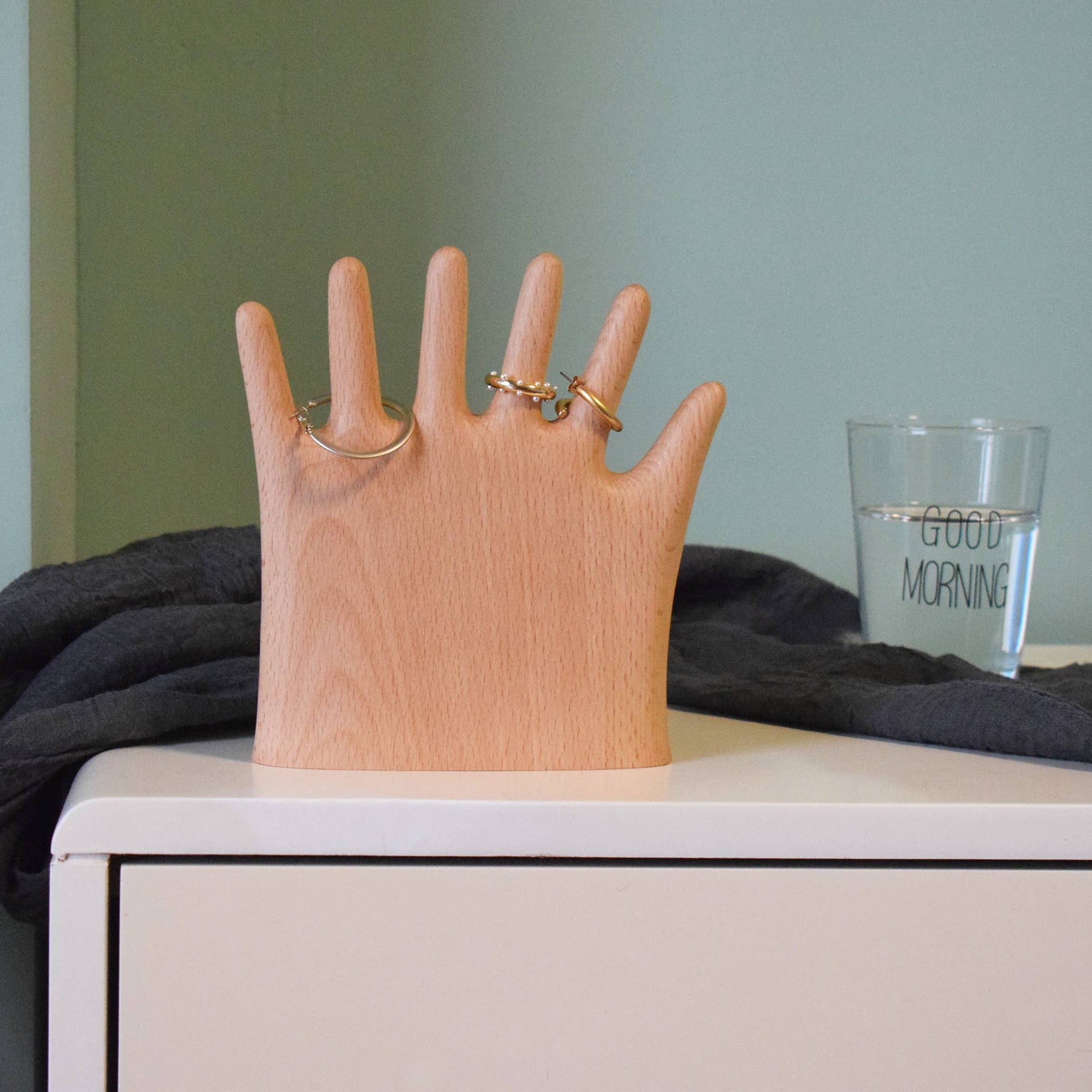 Wooden hand-shaped ring holder with rings on a surface with a glass of water in the background.