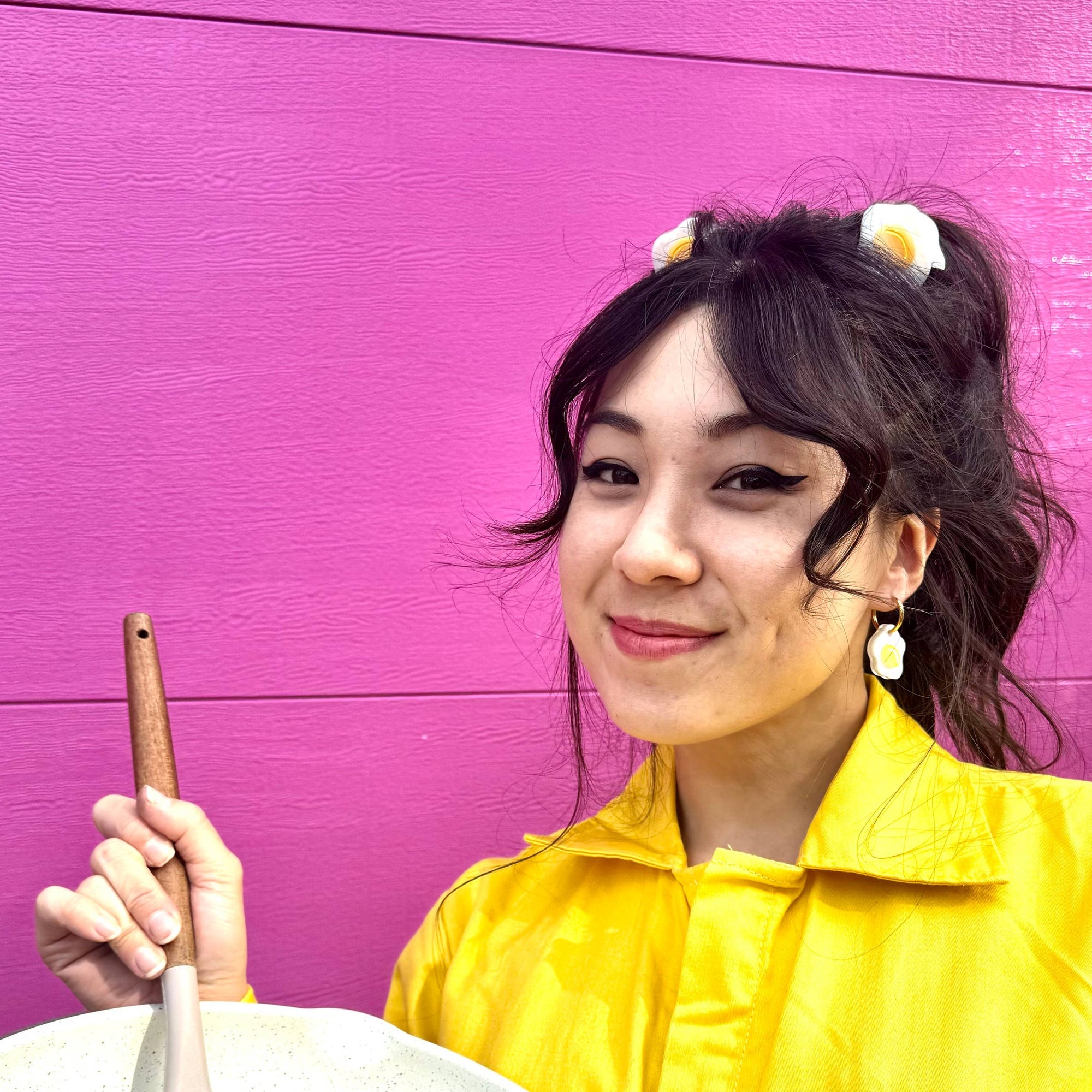 Person in a yellow raincoat holding a white frying pan with a spatula and egg against a purple background