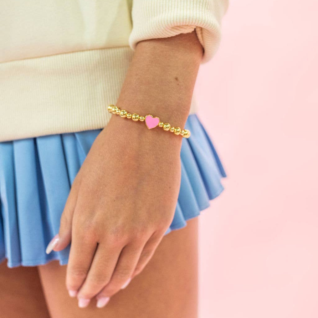 Hand wearing a gold bracelet with a pink heart charm on a pastel background