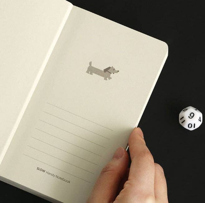Hand holding a notebook with a dachshund illustration next to a die on a black background