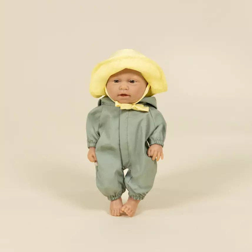 A baby doll or toy dressed in a sage green hooded jumpsuit with a yellow bow tie and a yellow sun hat, standing against a plain beige studio background.