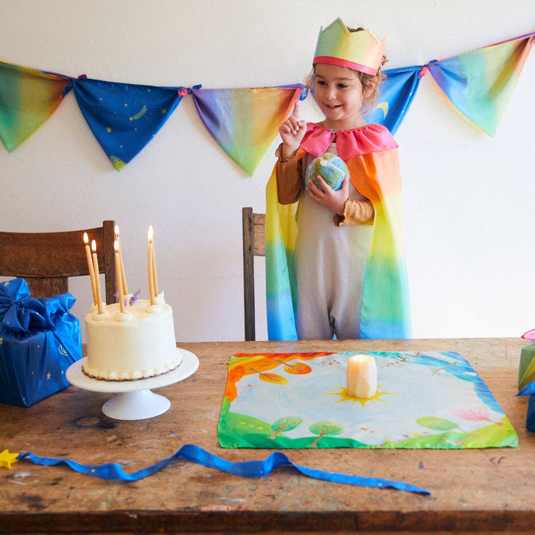 Child in a rainbow-themed costume standing behind a birthday cake with lit candles, surrounded by colorful decorations.