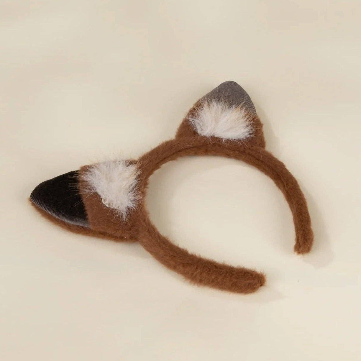 A brown fuzzy cat-ear headband with black-tipped ears and white inner-ear accents, photographed on a plain light beige background.