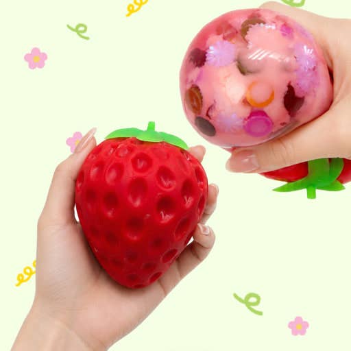Two hands holding strawberry-shaped squishy toys against a pale yellow background with small flower and script doodles. One toy is a red strawberry with green top, and the other is a pink translucent ball with colorful flower details and a red strawberry inside.