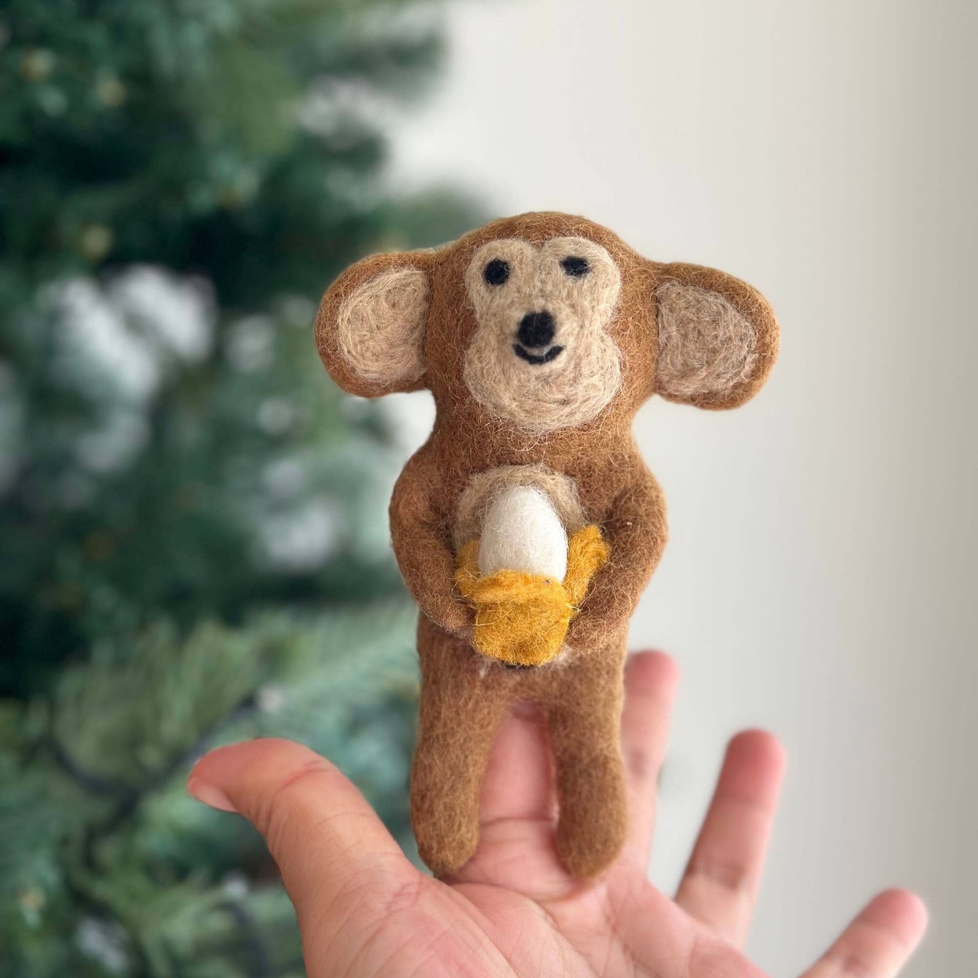 Hand holding a small brown monkey plush toy with a blurred Christmas tree in the background