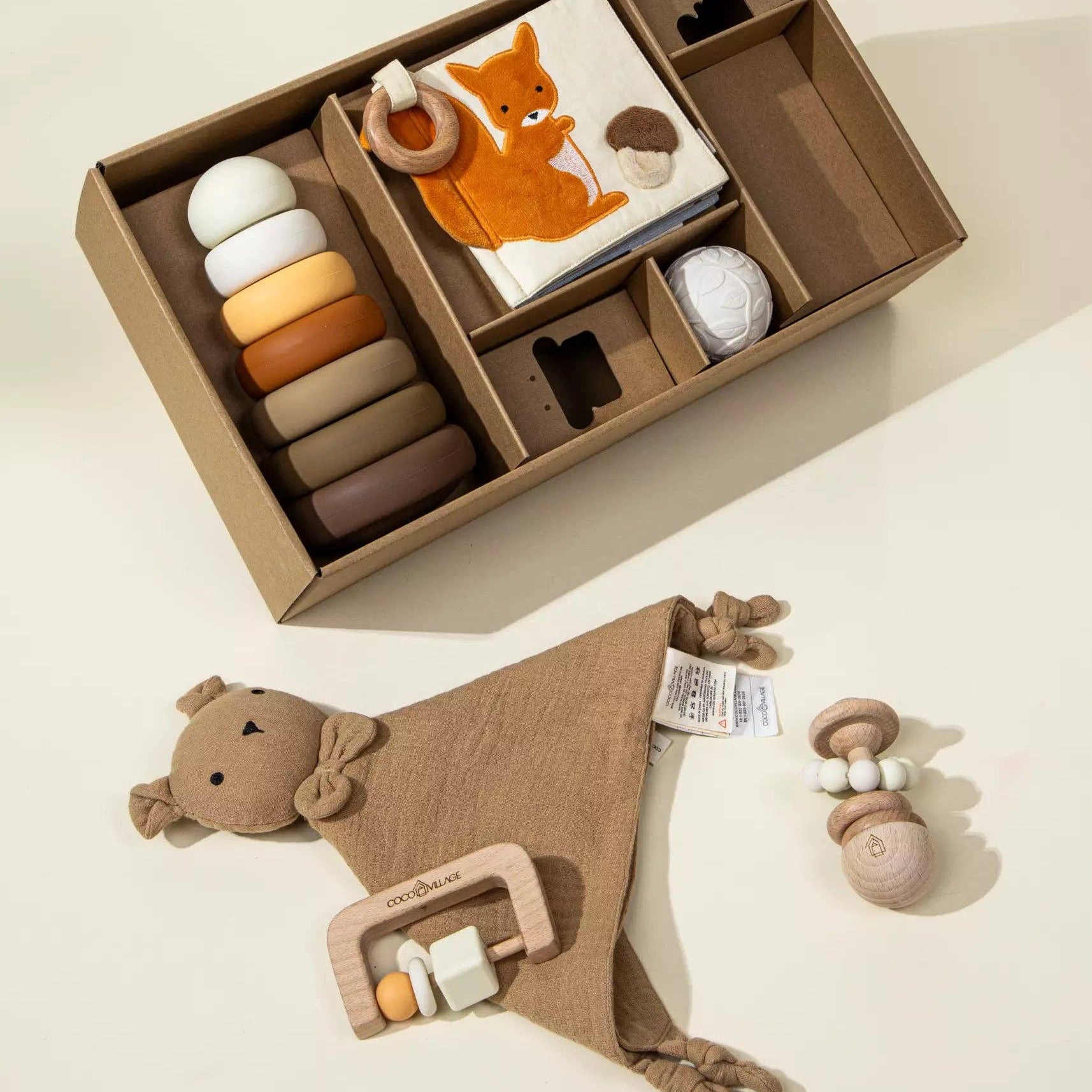 Baby toy set with wooden rings, teething ring, and soft blanket in a box on a light background