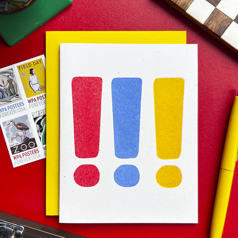 A white art print card featuring three large exclamation marks in red, blue, and yellow, layered over a bright yellow sheet on a textured red desk with vintage WPA posters and stationery props.