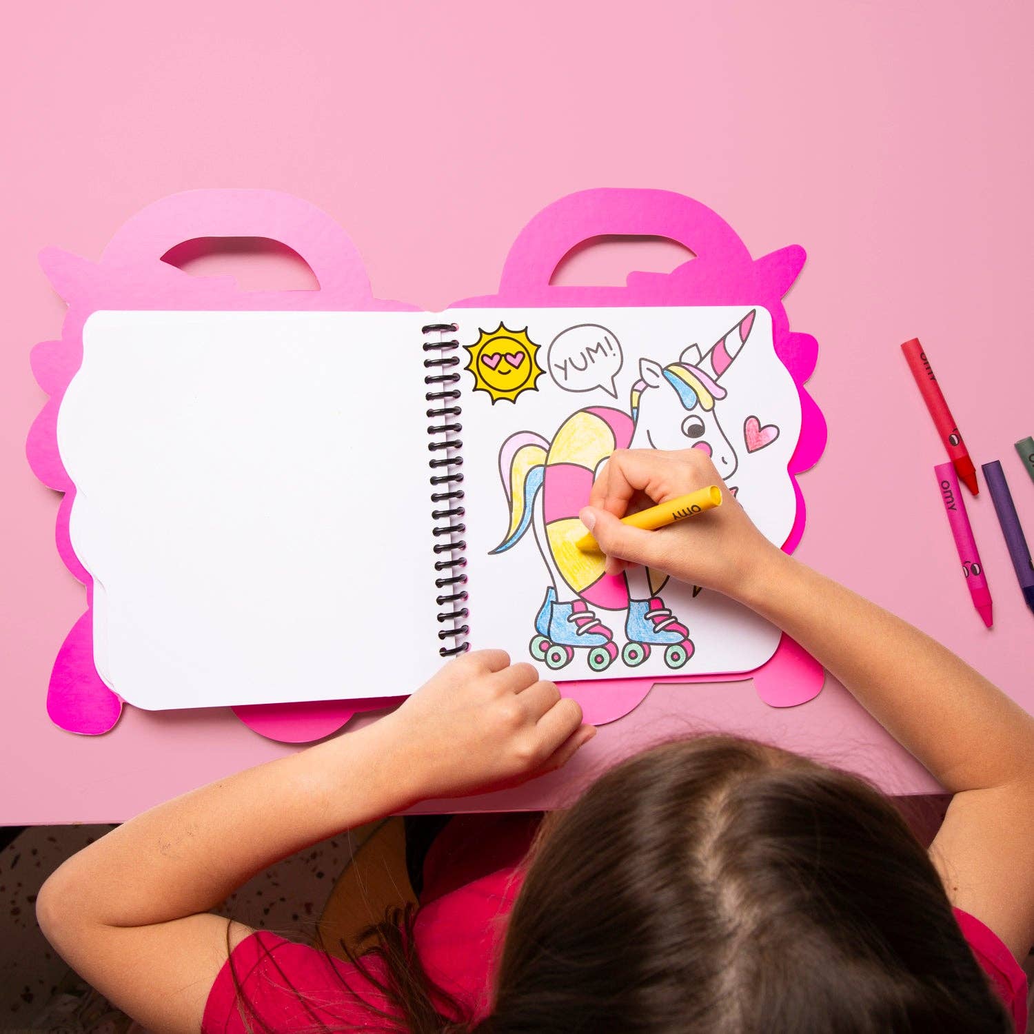Child coloring a unicorn on a pink desk with a coloring book and markers.