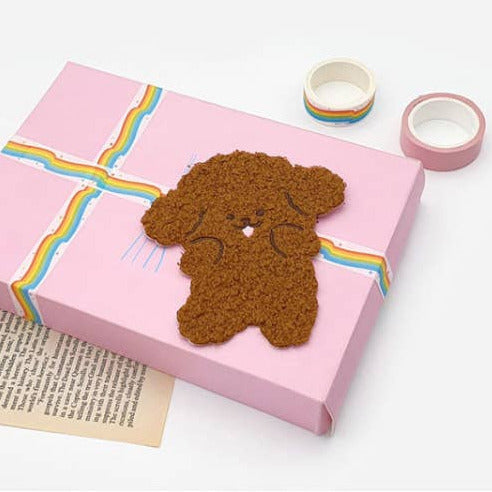 A pink gift box wrapped with a pastel rainbow ribbon and topped with a brown dog-shaped decorative item, styled on a white background with washi tape rolls and a printed paper sheet.