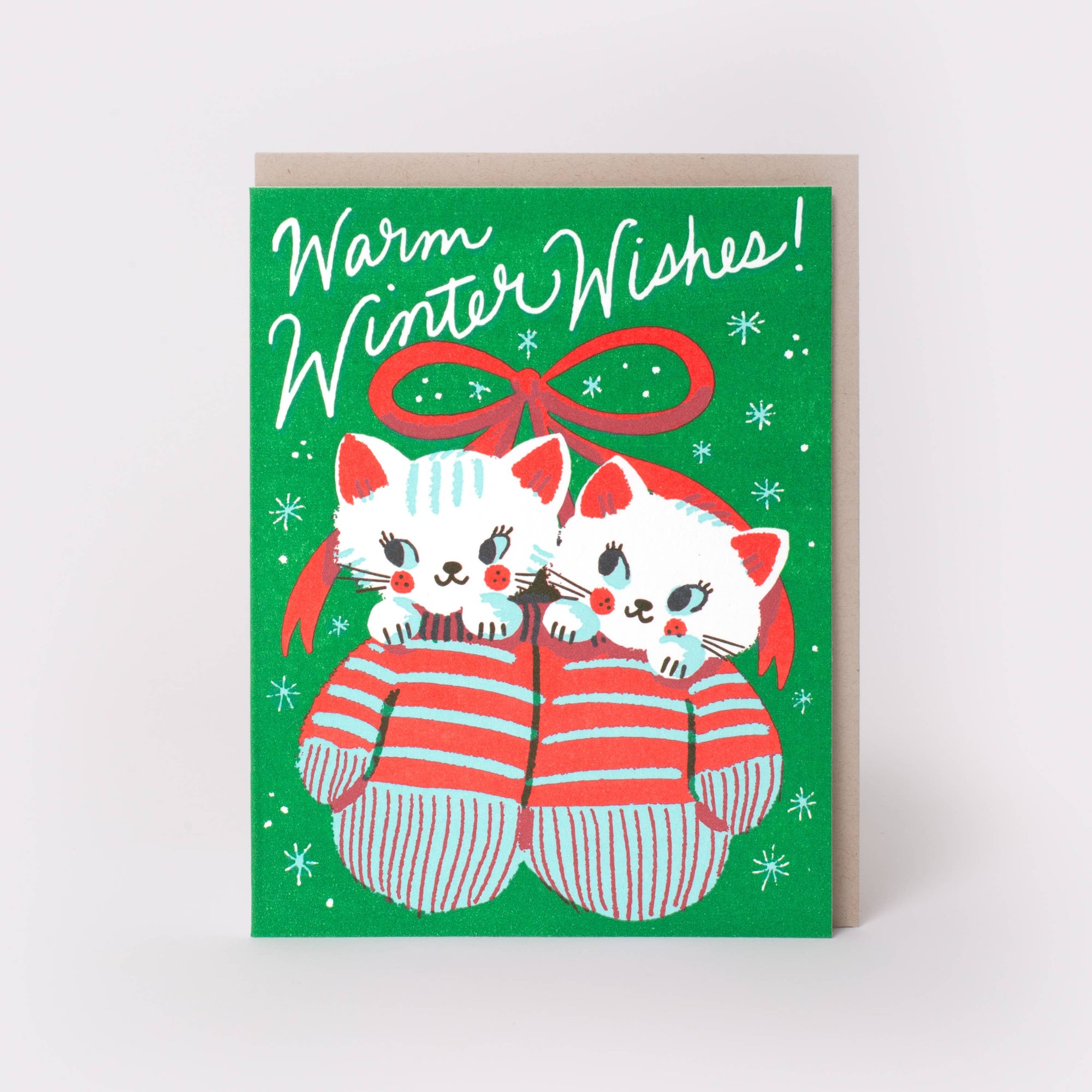 A Christmas greeting card with a green background, white script text reading 'Warm Winter Wishes!', and an illustration of two white kittens wearing red-and-white striped socks under a red bow. A plain light brown envelope is partially visible behind the card.