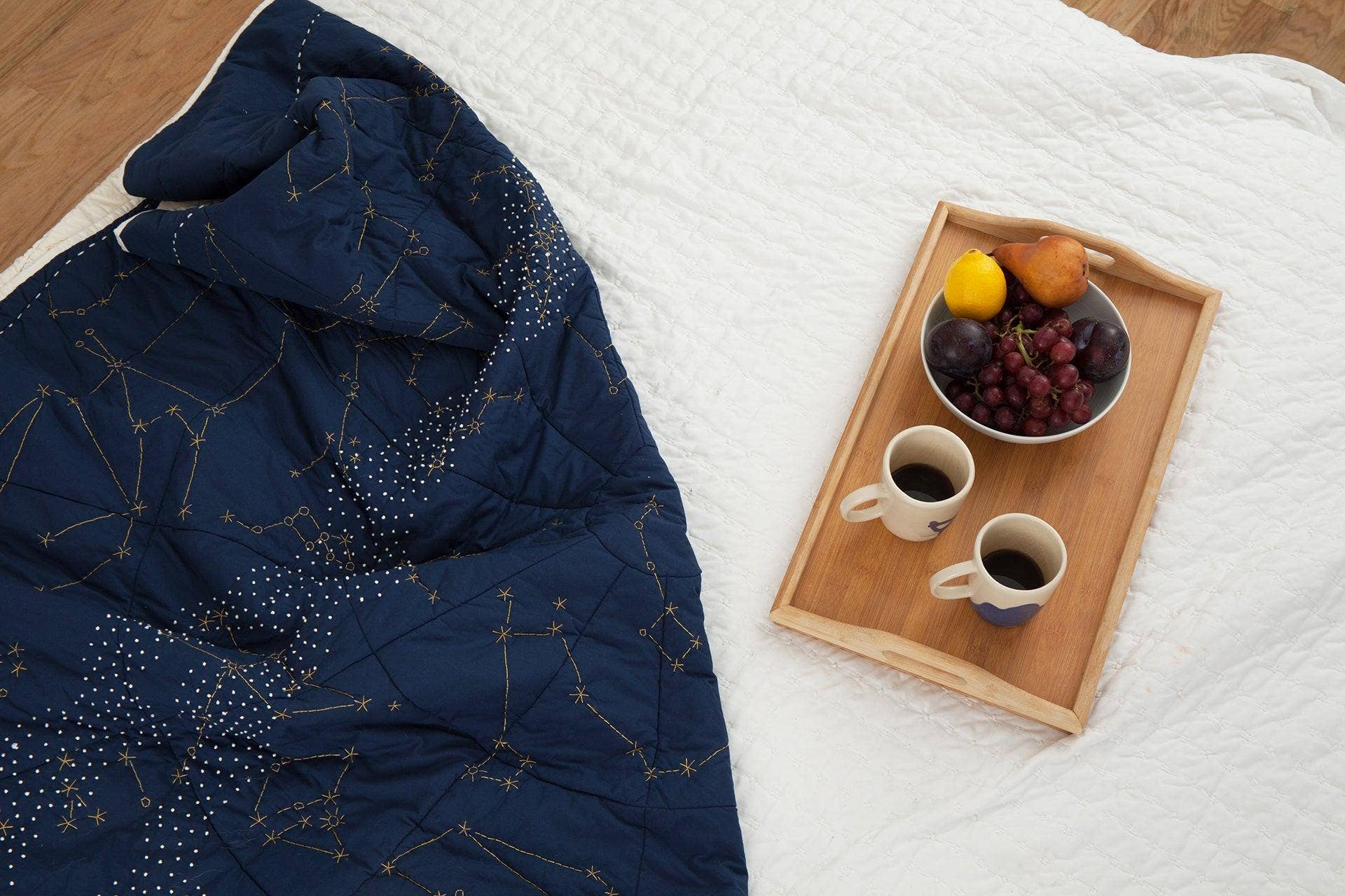 Navy blue quilt with star pattern on a white bed, next to a wooden tray with coffee and fruit.