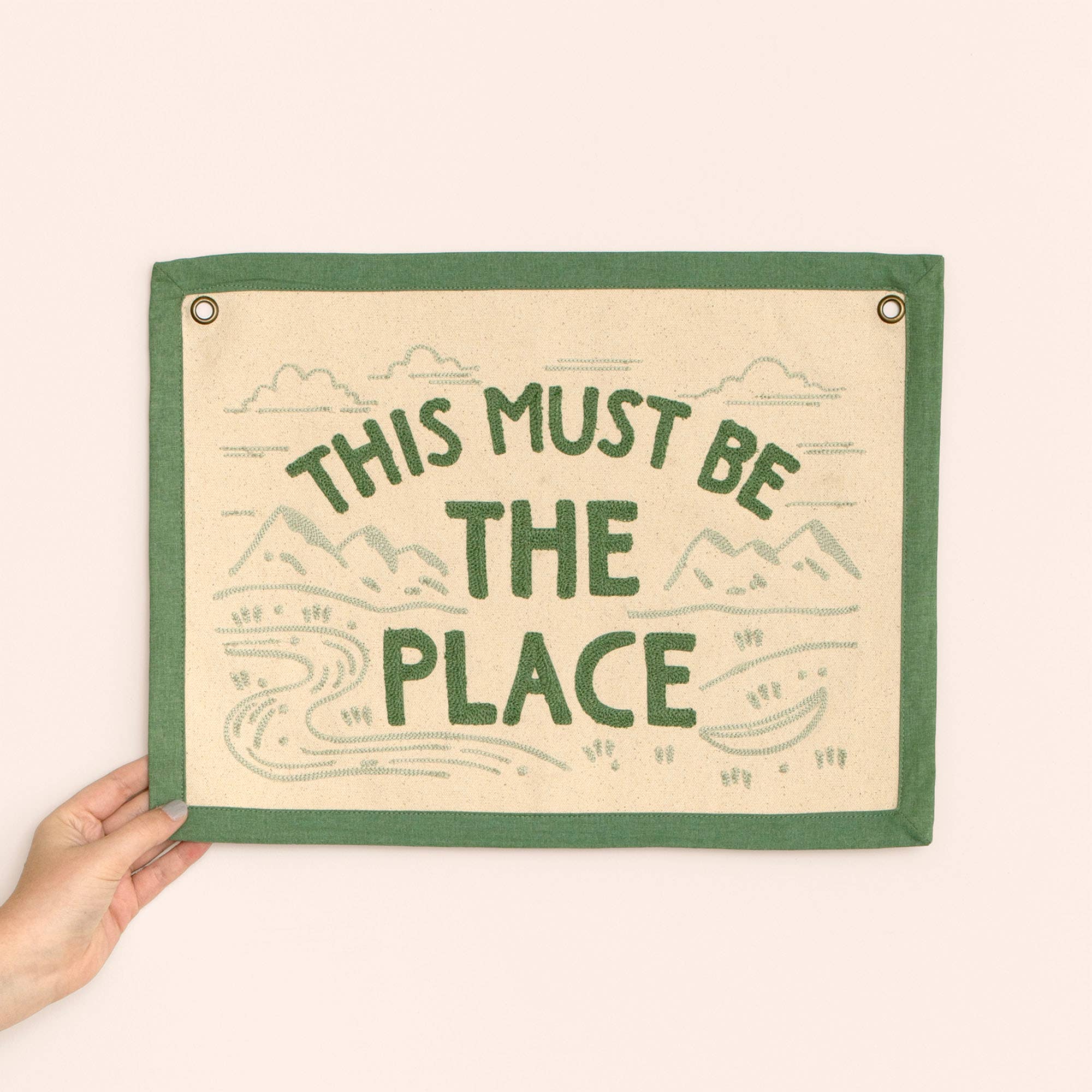 Fabric sign with 'This Must Be The Place' text held by a hand on a white background