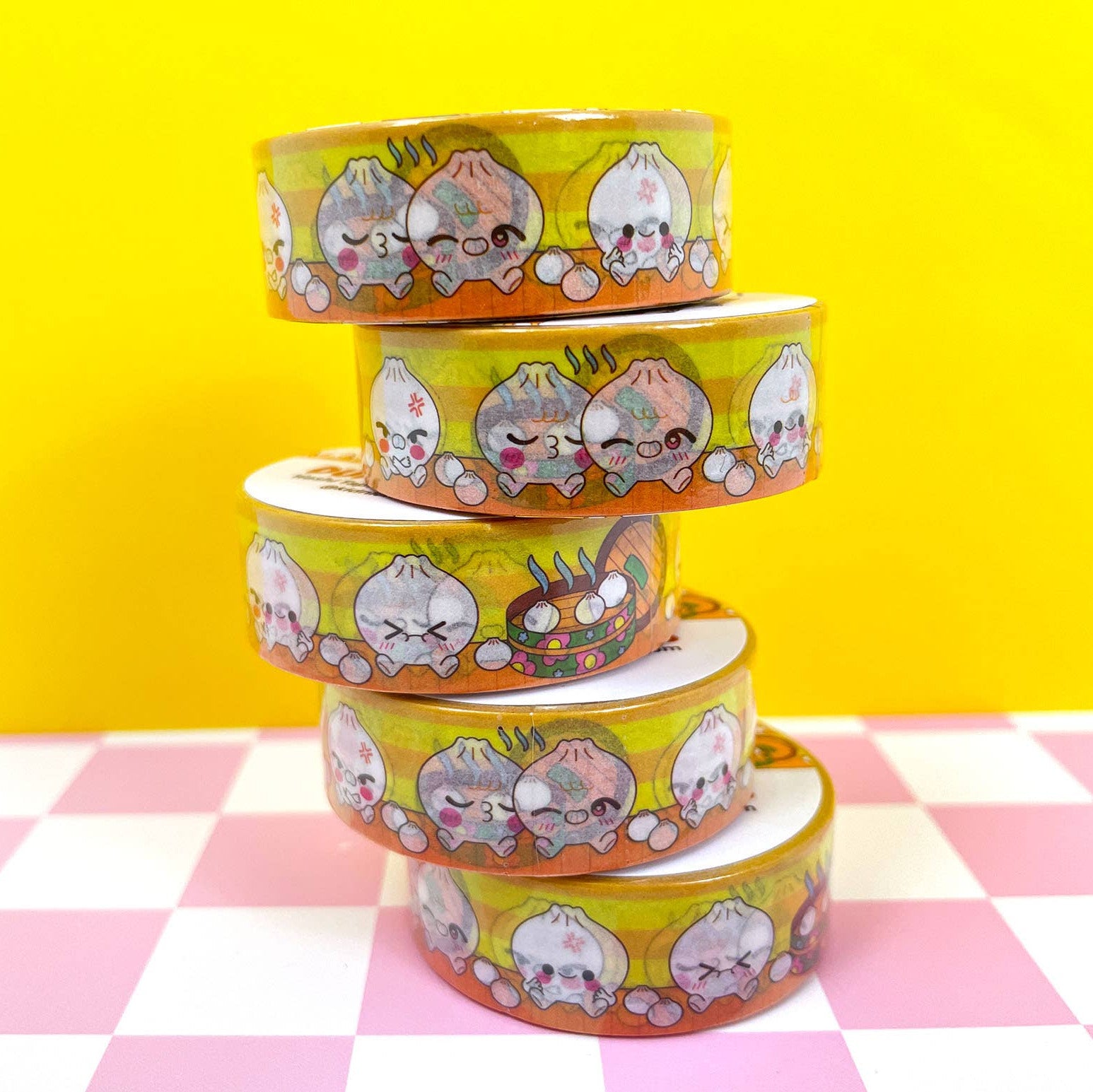 Stack of colorful washi tape rolls with cartoon designs on a pink and white checkered tablecloth against a yellow background.