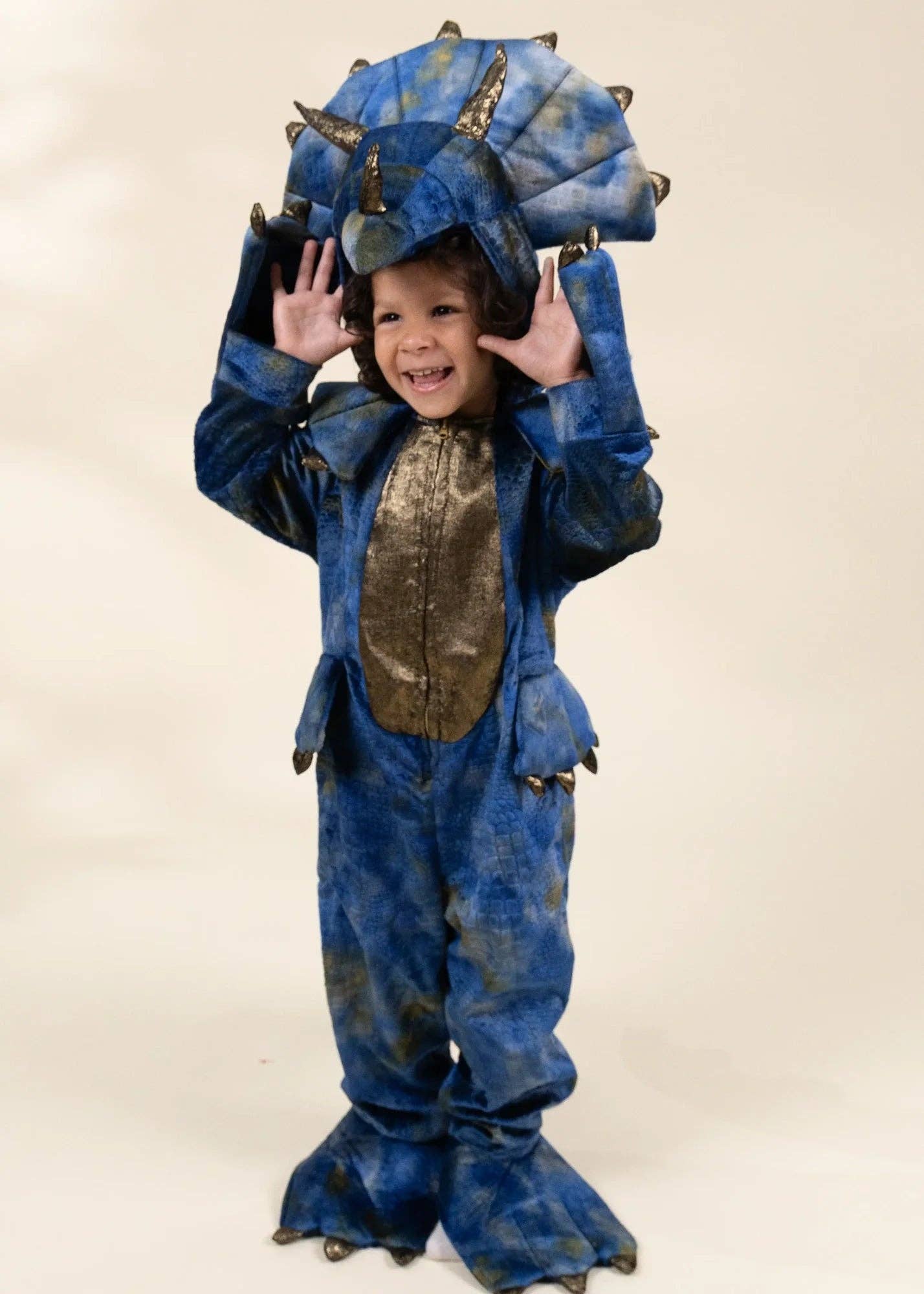 A young child wearing a blue dinosaur costume with a large spiky head crest, textured blue body, and metallic bronze chest panel, standing against a plain beige studio background.