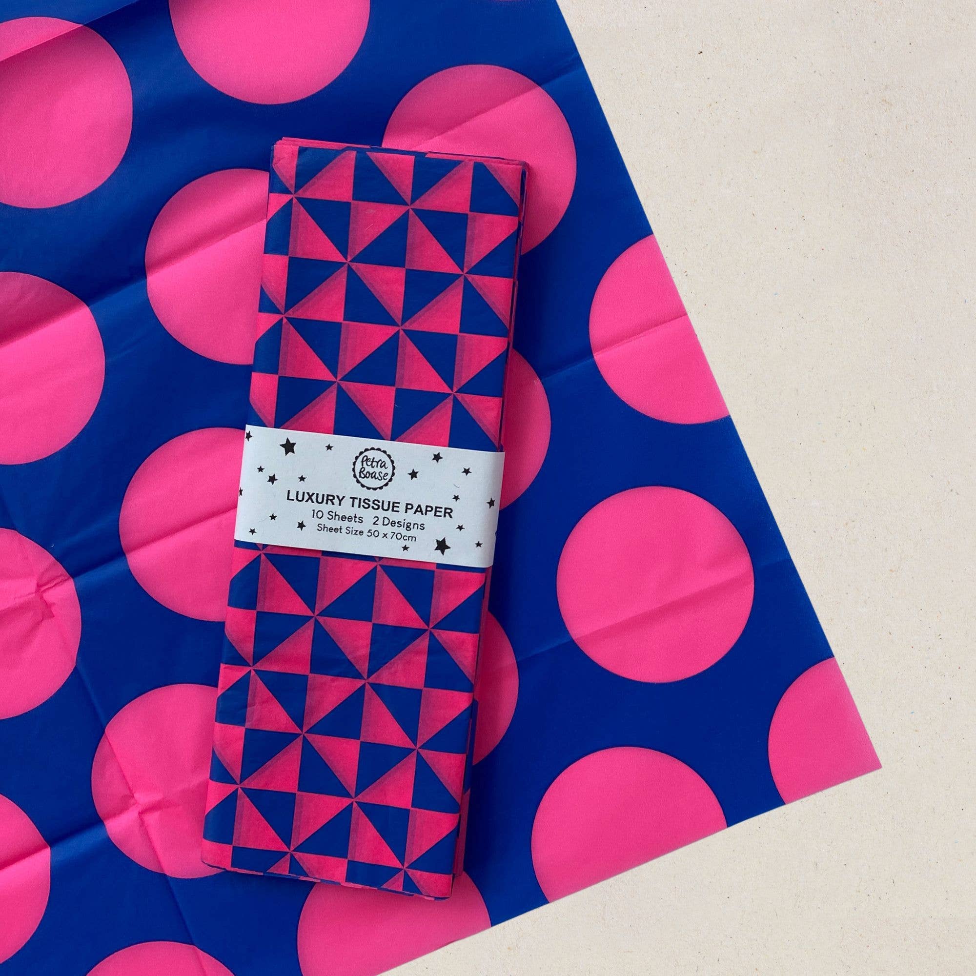 A roll of patterned luxury tissue paper wrapped with a white paper band, placed diagonally on top of a matching blue and pink patterned sheet. The band reads 'The Paper Club LUXURY TISSUE PAPER'.