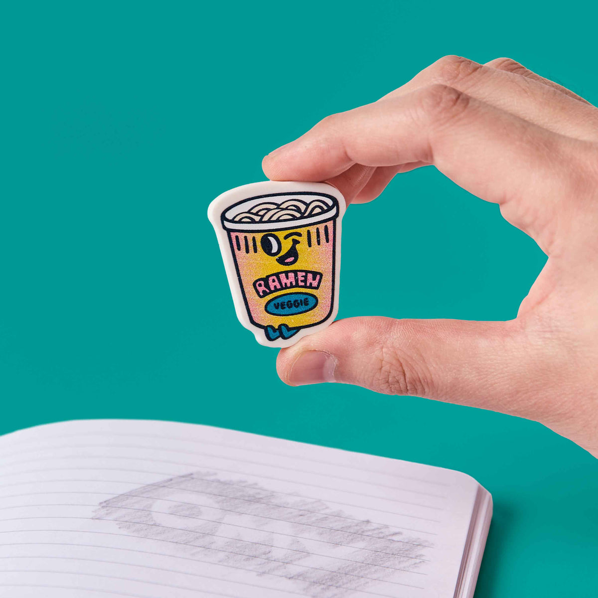 Hand holding a cartoon ramen noodle cup sticker above an open notebook with a teal background