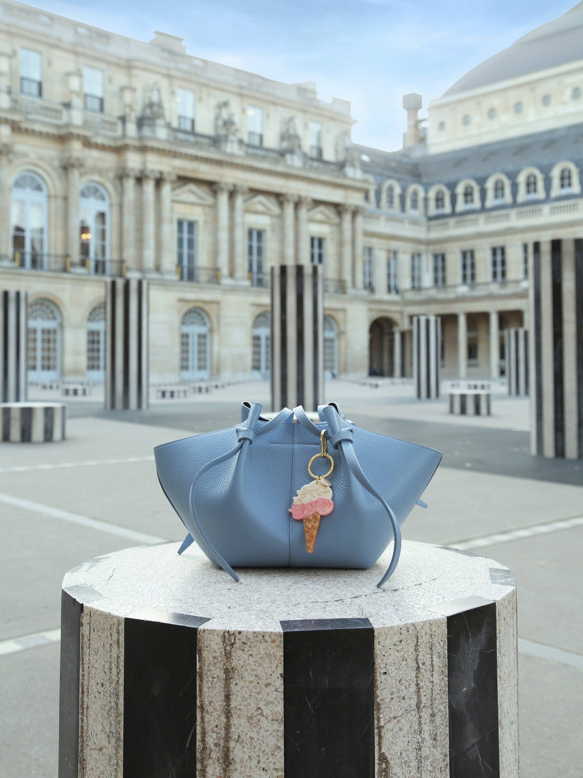 Light blue handbag with a pink ice cream charm on a pedestal in front of a classical building.