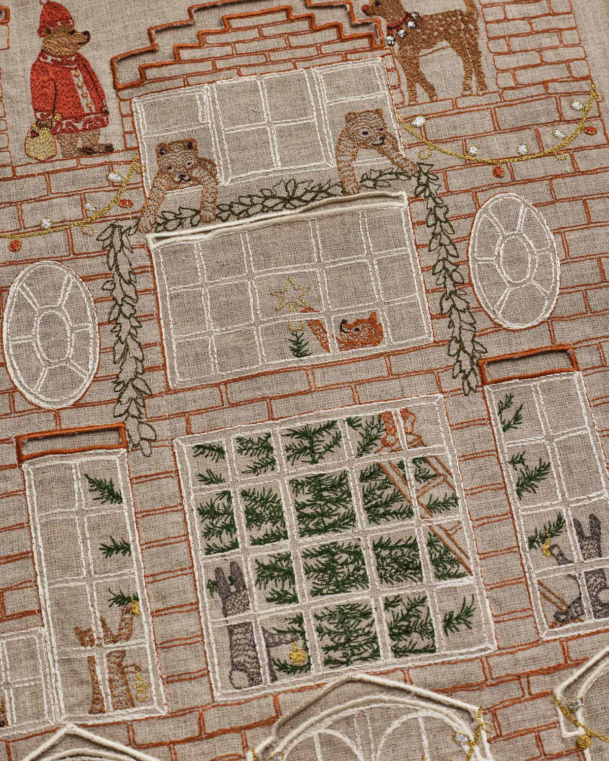 Close-up of an embroidered textile featuring a brick house facade with multiple windows, green garlands, stockings, and small illustrated animals including a bear in a red coat and a dog.