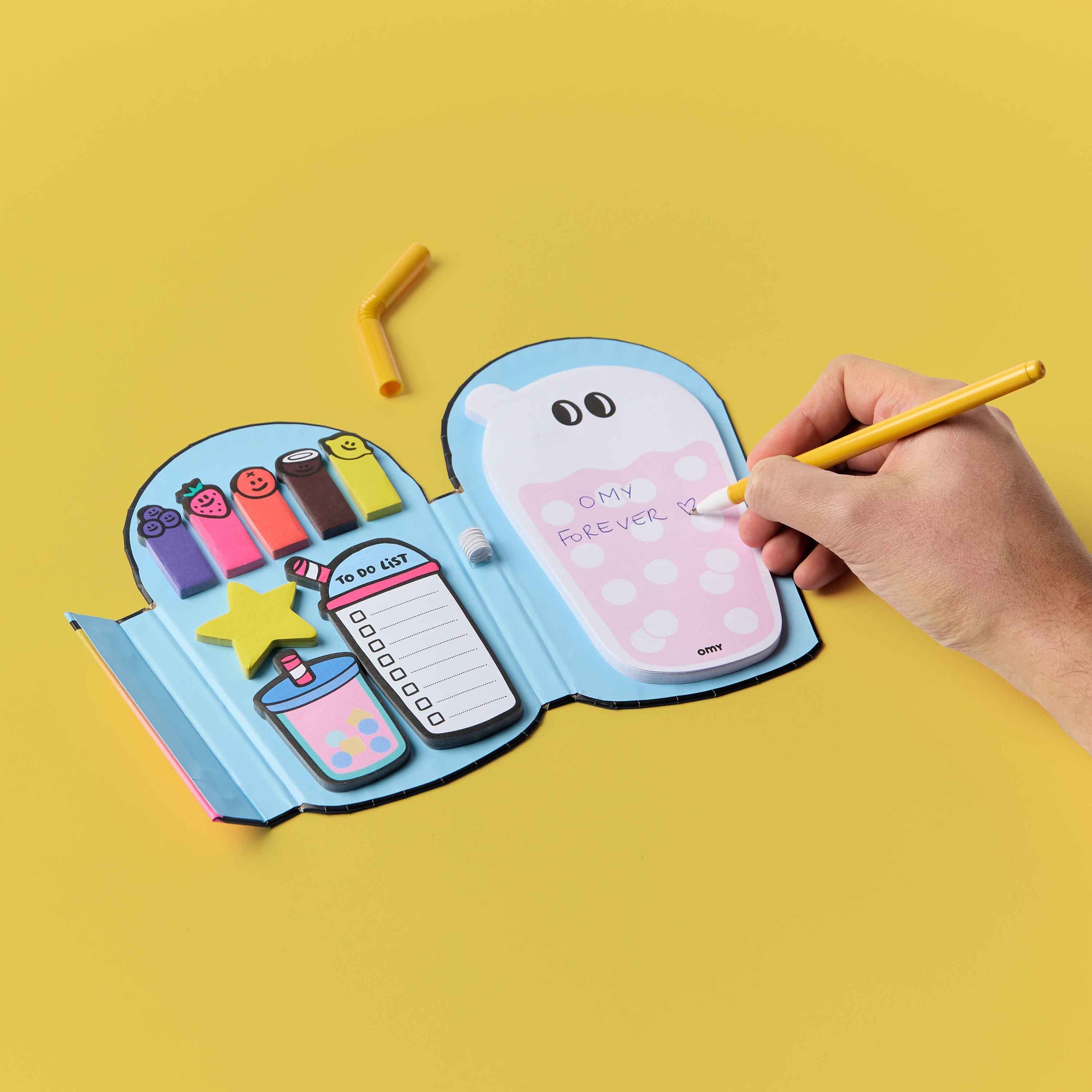 Hand holding a yellow pen over an open notebook with colorful illustrations on a yellow background