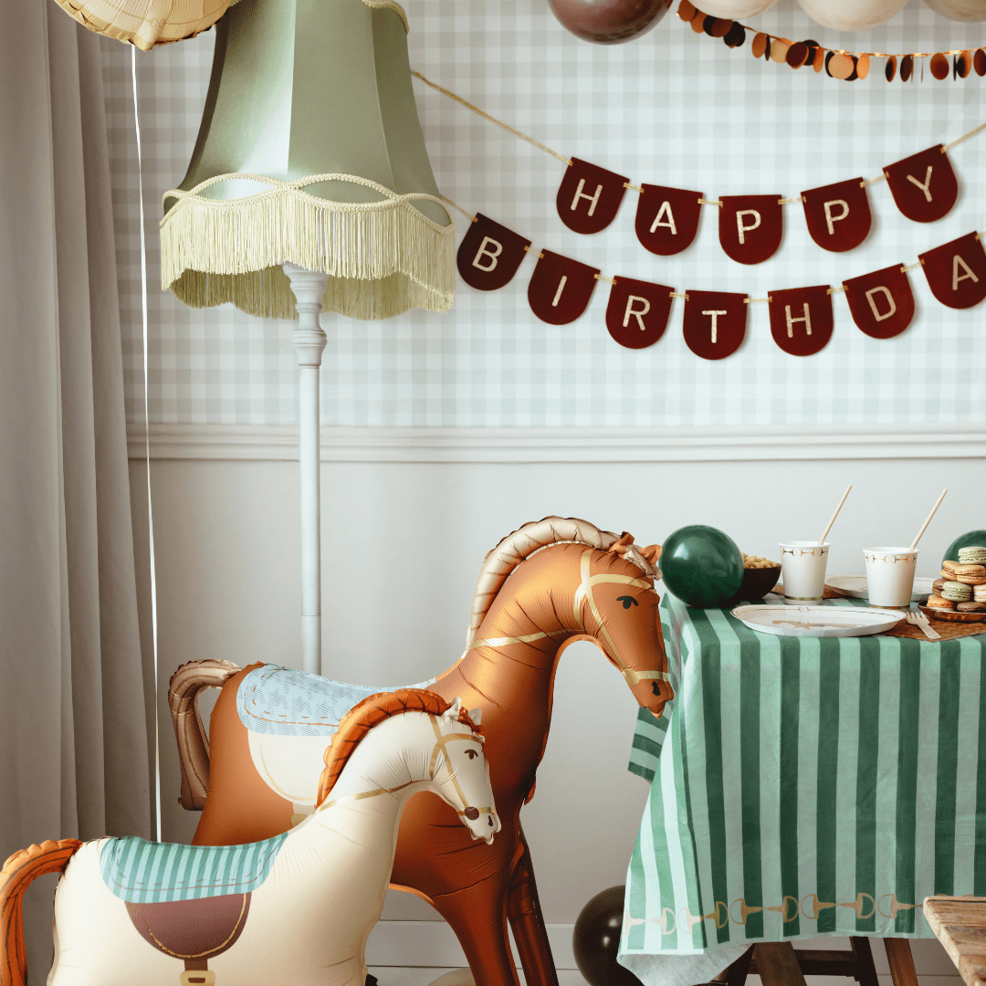 A styled birthday party scene with a green and white striped tablecloth, a green and white checkered wall, a hanging 'HAPPY BIRTHDAY' banner, a vintage-style floor lamp, and two toy horses in the foreground.