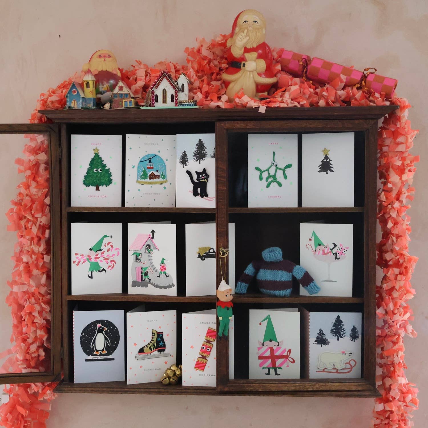 Wooden shelf with Christmas-themed cards and decorative items against a pink background