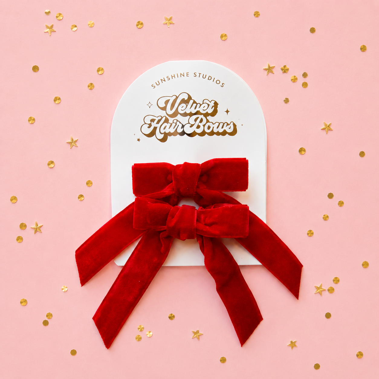 A red velvet hair bow attached to a white display card printed with the brand name Sunshine Studios and the product name Velvet Hair Bow, set against a pink background with scattered gold confetti.