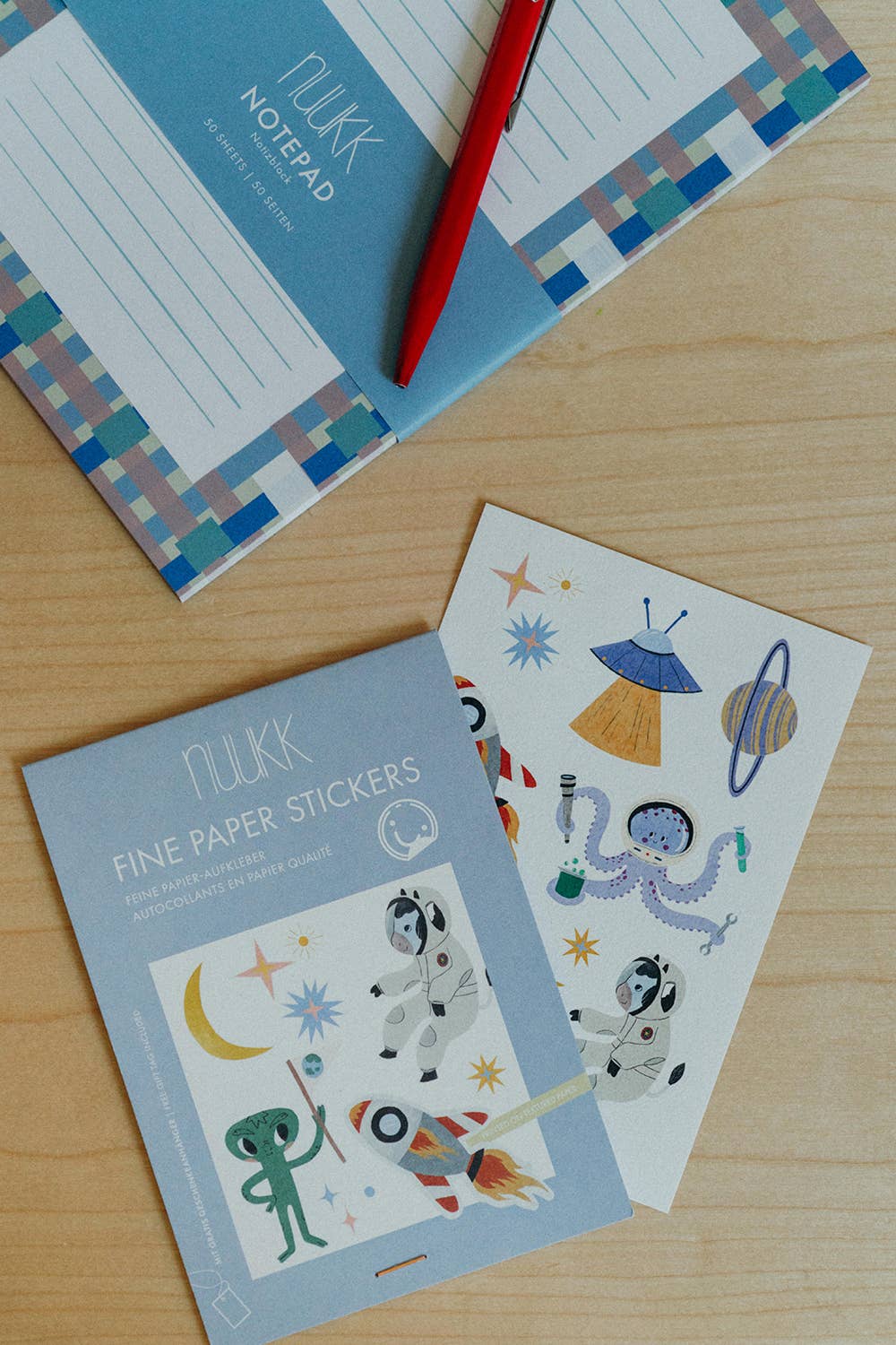 Nukk fine paper stickers with an outer space theme, displayed on a light wood table next to a matching blue notepad and a red pen.