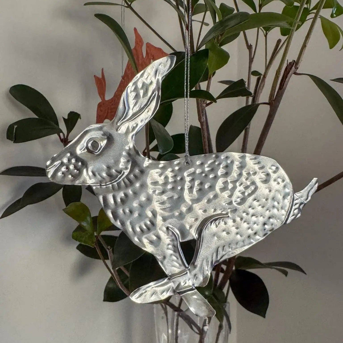 Decorative metal rabbit in a glass vase with greenery against a plain wall.