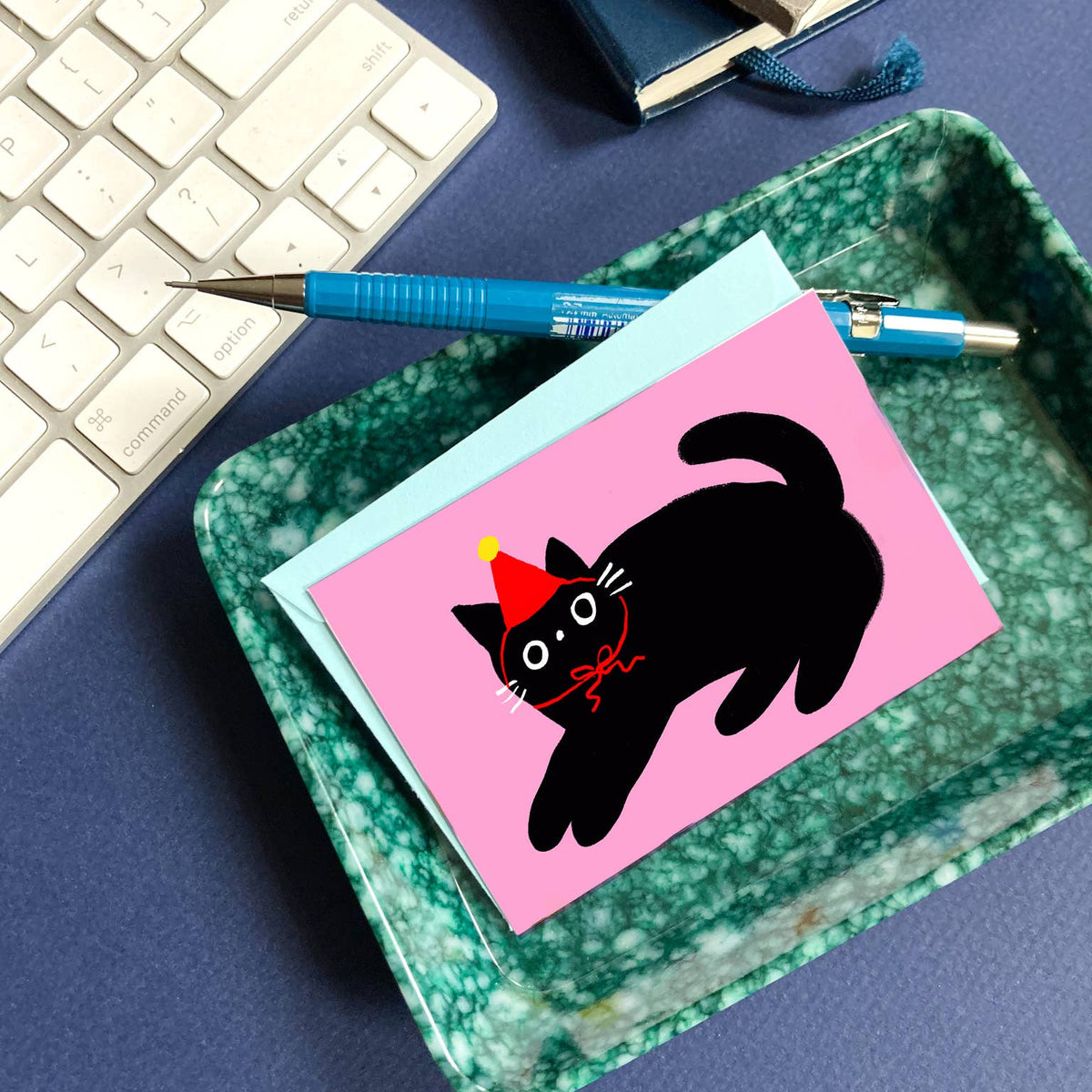 card with a black cat illustration on pink paper, placed on a green marble surface with a keyboard in the background.