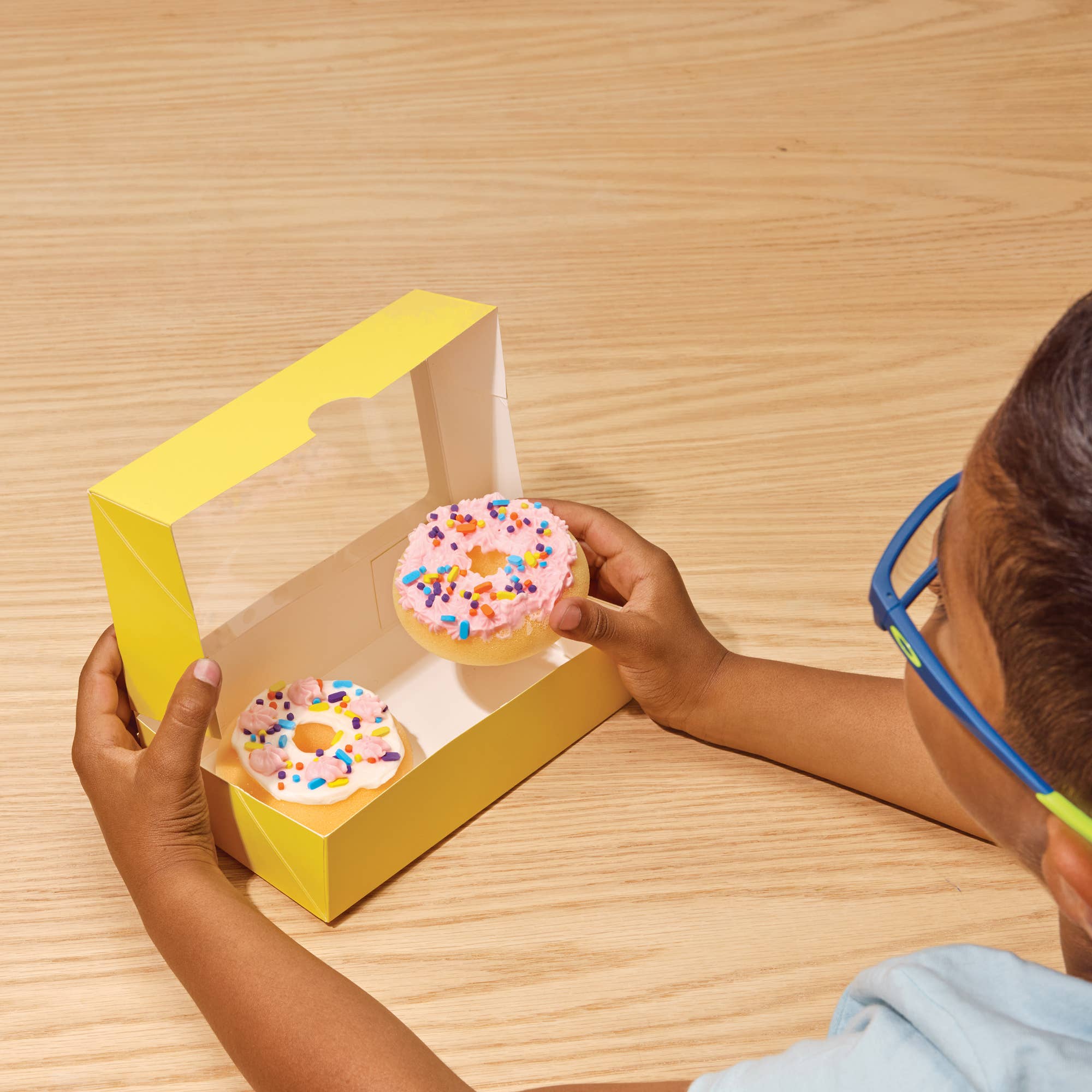 Child holding a pink donut with sprinkles out of a yellow box on a wooden table
