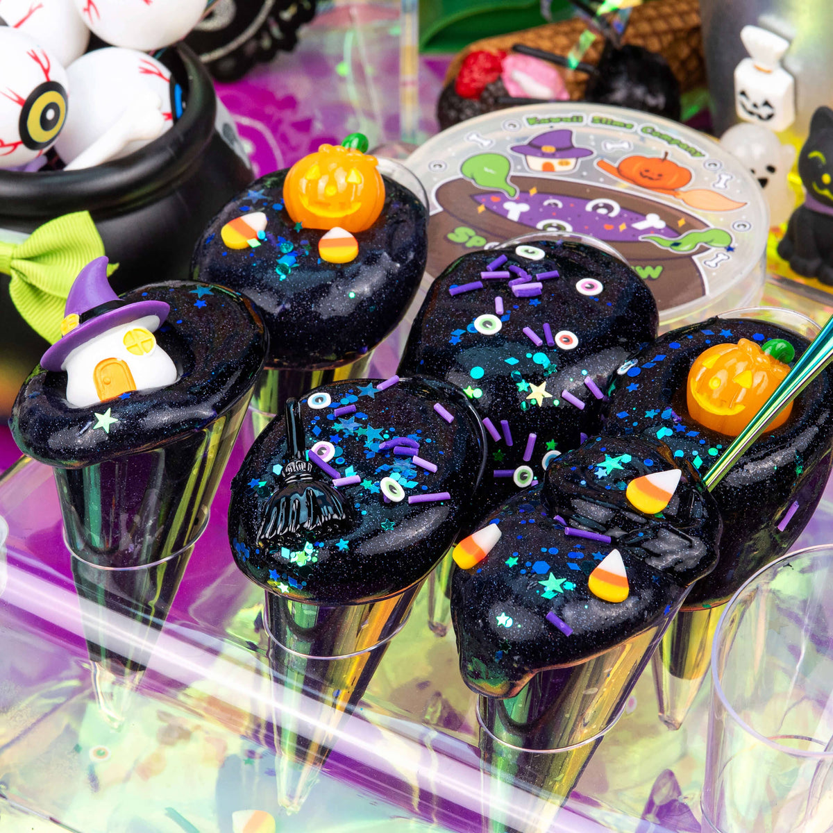 Halloween-themed star-shaped lollipops with black glittery bodies, purple sprinkles, orange pumpkin toppers, candy corn accents, and small witch motifs, displayed in a colorful festive setting.