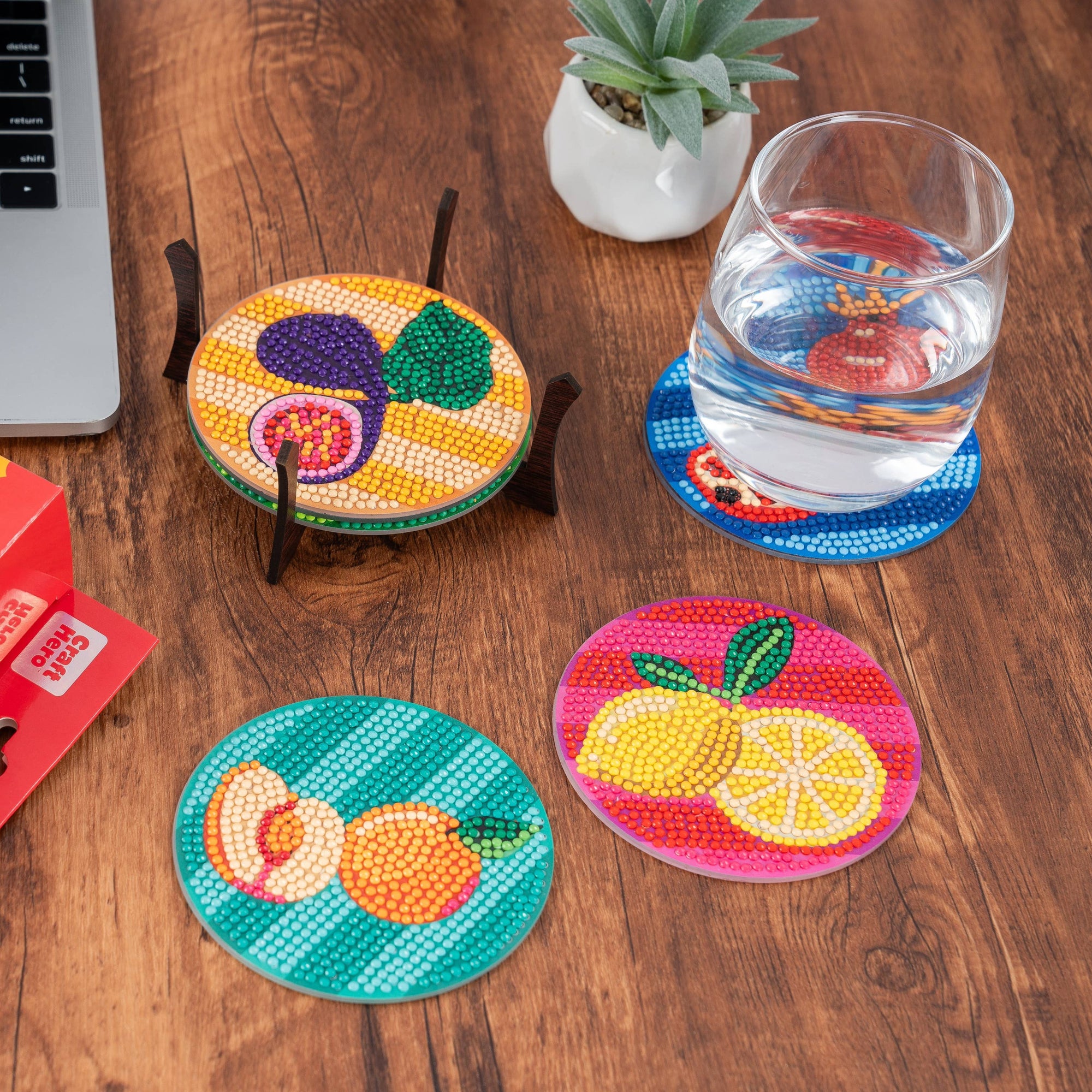 Colorful coasters with fruit designs on a wooden table next to a laptop and glass of water.