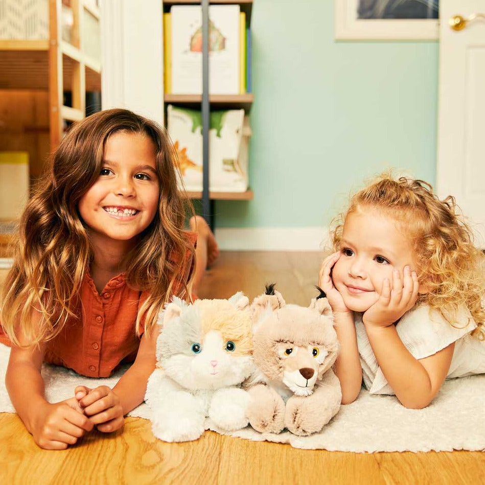 Two young girls lying on a wooden floor with stuffed animals in a room with bookshelves.