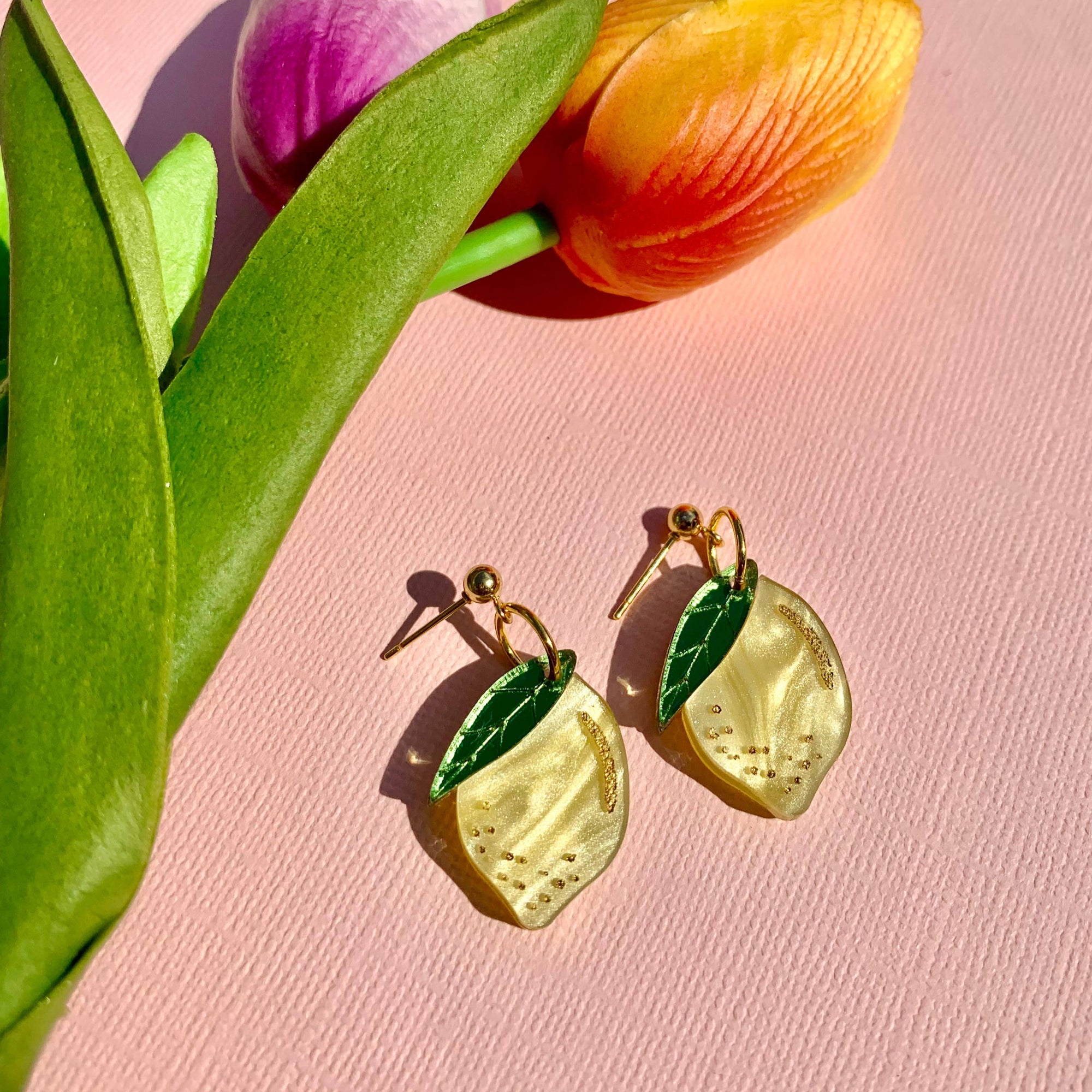 Lemon-shaped earrings with green leaves on a pink background with tulips.