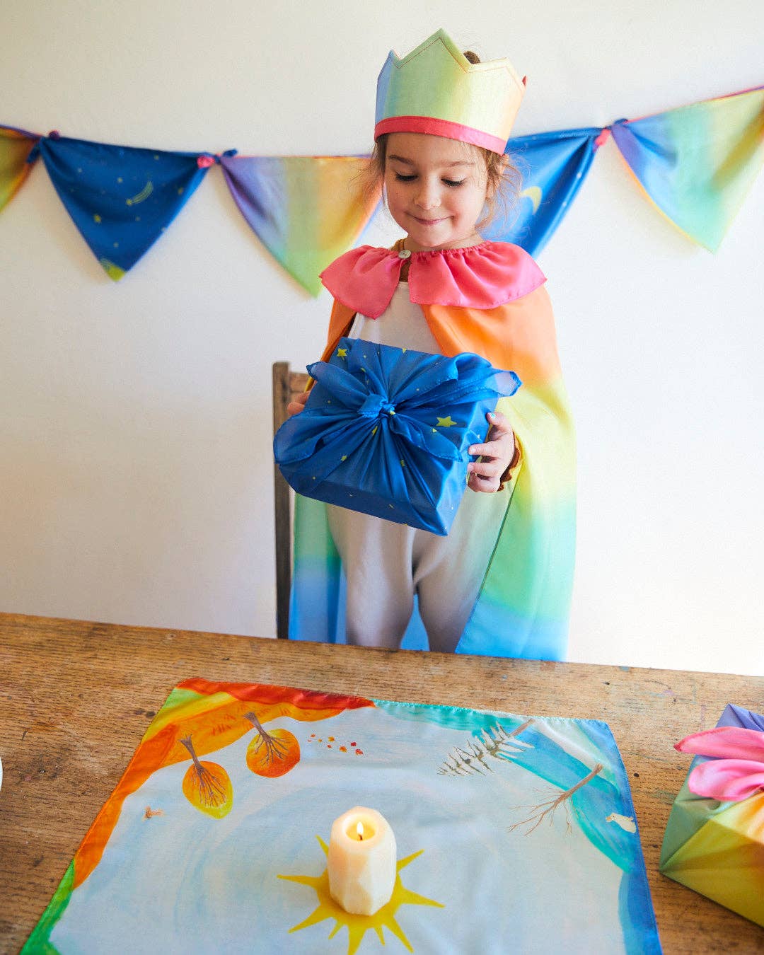Child in a rainbow costume holding a blue gift box with colorful decorations and a candle on a table.