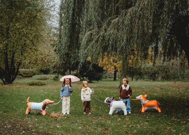 Three children standing in a park with a weeping willow tree, each holding a colorful dog-shaped toy on a leash. The toys resemble a dachshund, a white and black dog, and an orange fox.