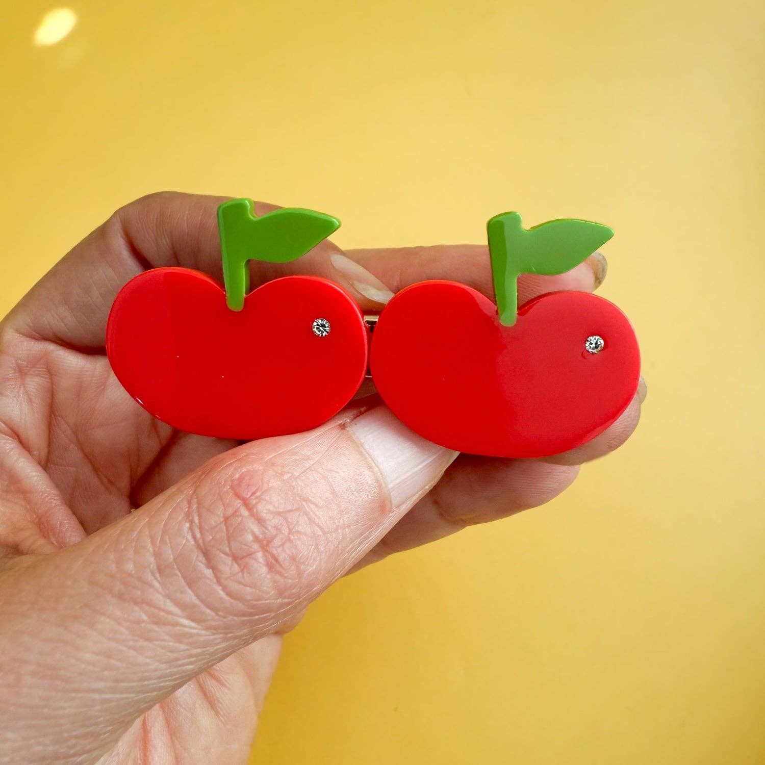 Red apple-shaped earrings with green leaves held by a hand against a yellow background