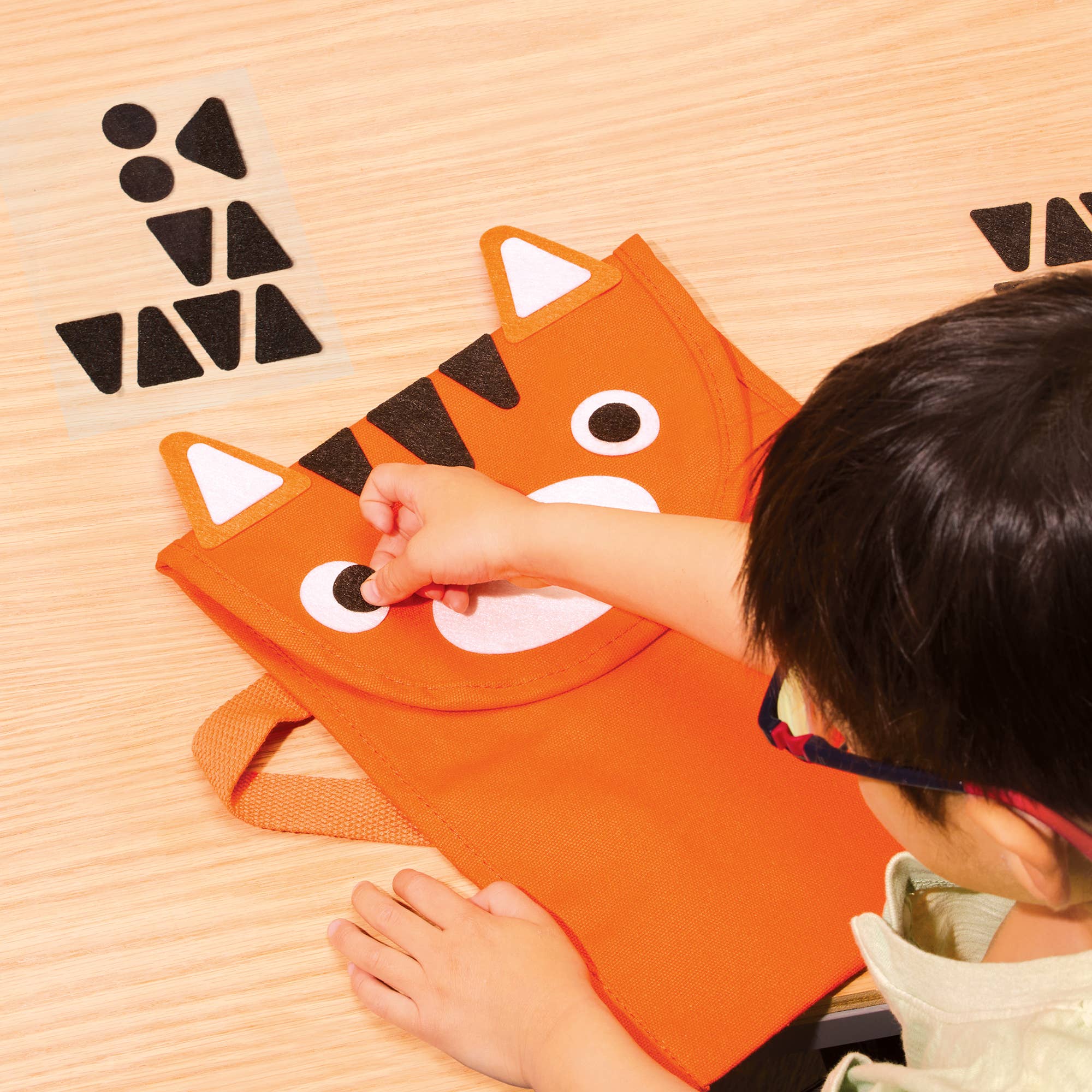 A child's hands and head are visible as they assemble a felt orange animal face craft on a light wood tabletop. The craft features a fox or cat design with white and black felt pieces, and other black geometric felt pieces are laid out nearby.