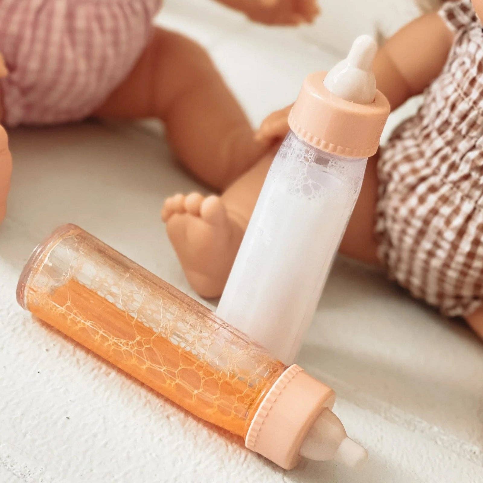 Baby bottle and bottle with textured orange design on a light surface, with dolls in the background.