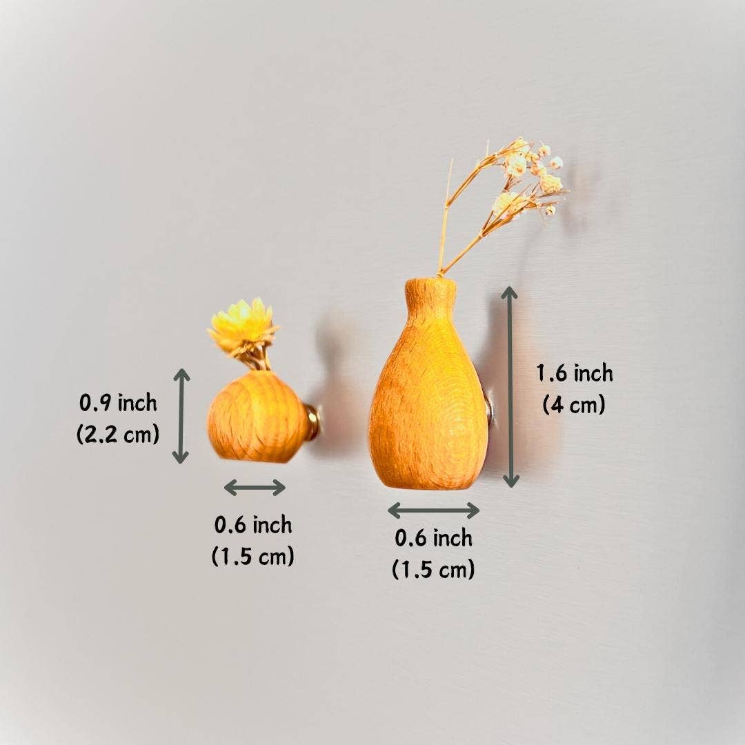 Two small dried flower vases shown against a plain light gray background, with measurement arrows and text indicating the height and width of each piece. One vase is yellow-orange with a small yellow flower, and the other is tan-orange with a slightly larger dried stem.