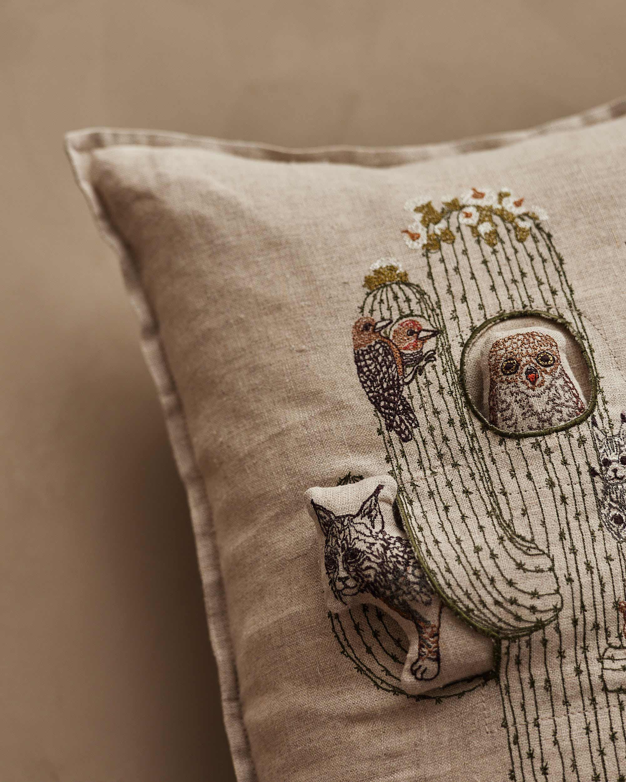 A close-up of a beige linen throw pillow featuring embroidered desert animals, including a cactus, owl, fox, and bird, in muted earthy tones against a matching beige background.