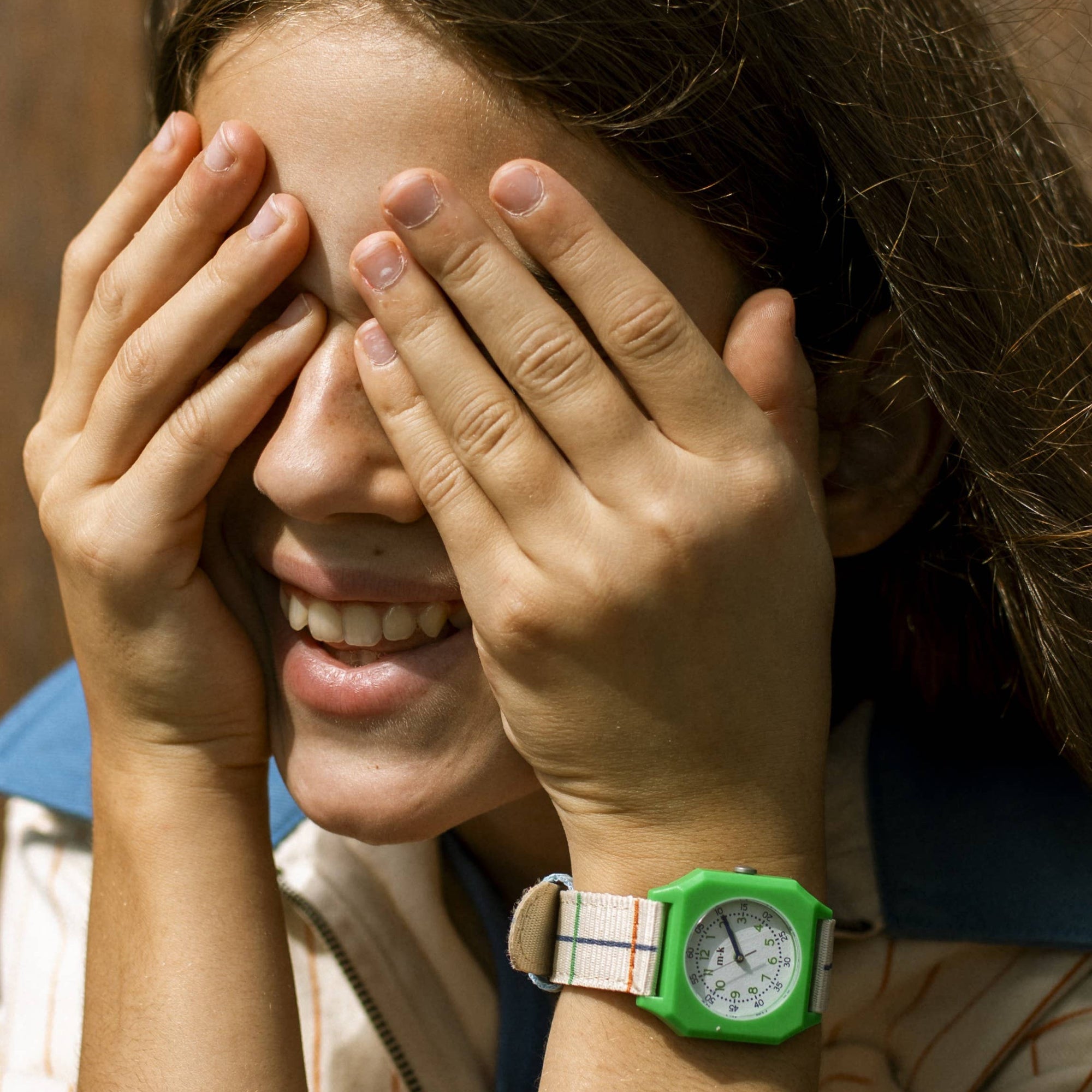Person wearing a green watch with a patterned strap, covering their face with their hands.