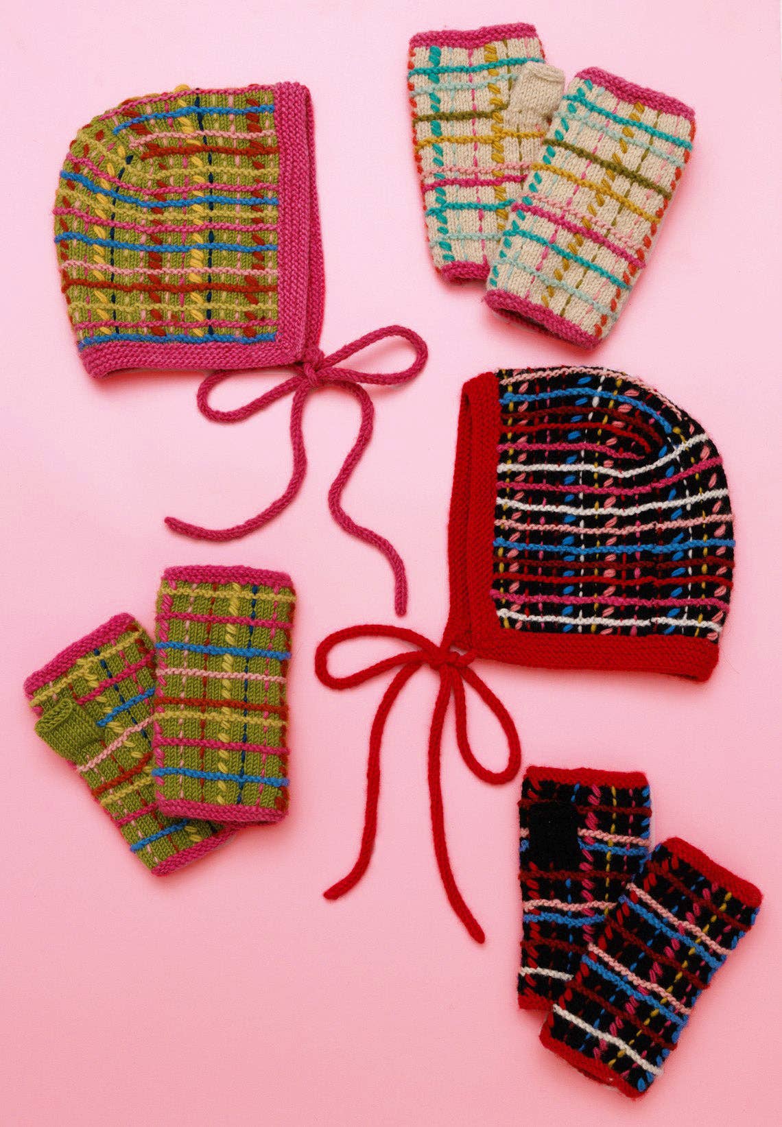 A set of hand-knitted baby accessories including bonnets and finger mittens in multicolored plaid yarn, arranged on a solid pink background.