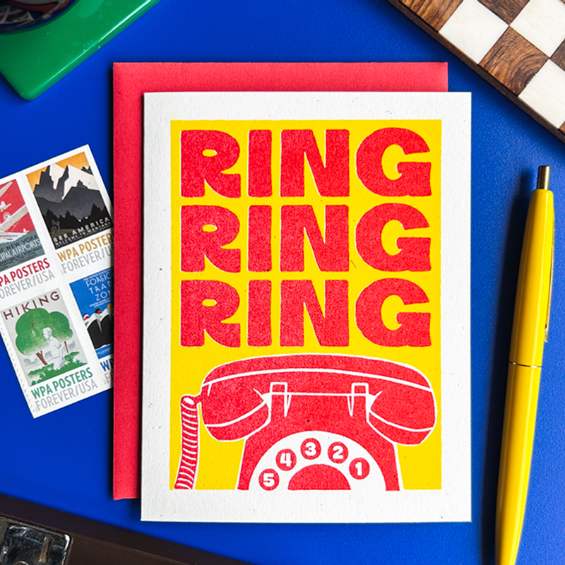 A square art print with a white border featuring the word 'RING' repeated in bold red letters above a red rotary phone illustration on a yellow background. The print is displayed on a blue desk with a red envelope behind it, surrounded by small postcards, a yellow pen, and other desk items.