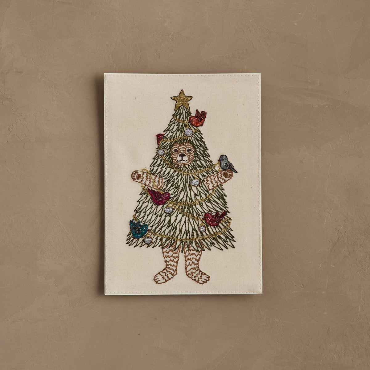 A square cream-colored embroidered art piece featuring a teddy bear dressed as a Christmas tree, with a gold star on top, decorated ornaments, and small birds on the branches, displayed against a textured brown background.