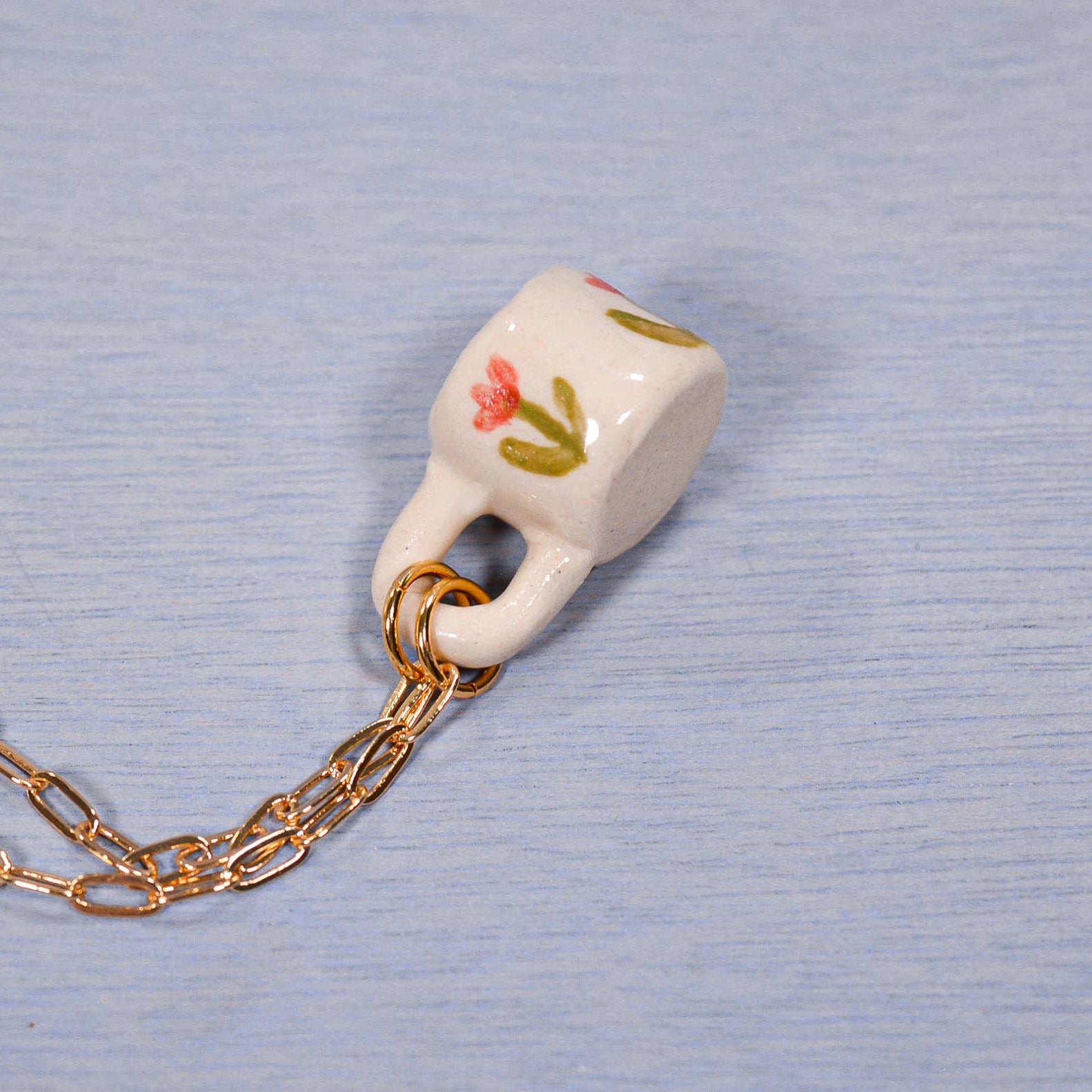 Small ceramic mug charm on a gold chain against a light blue background