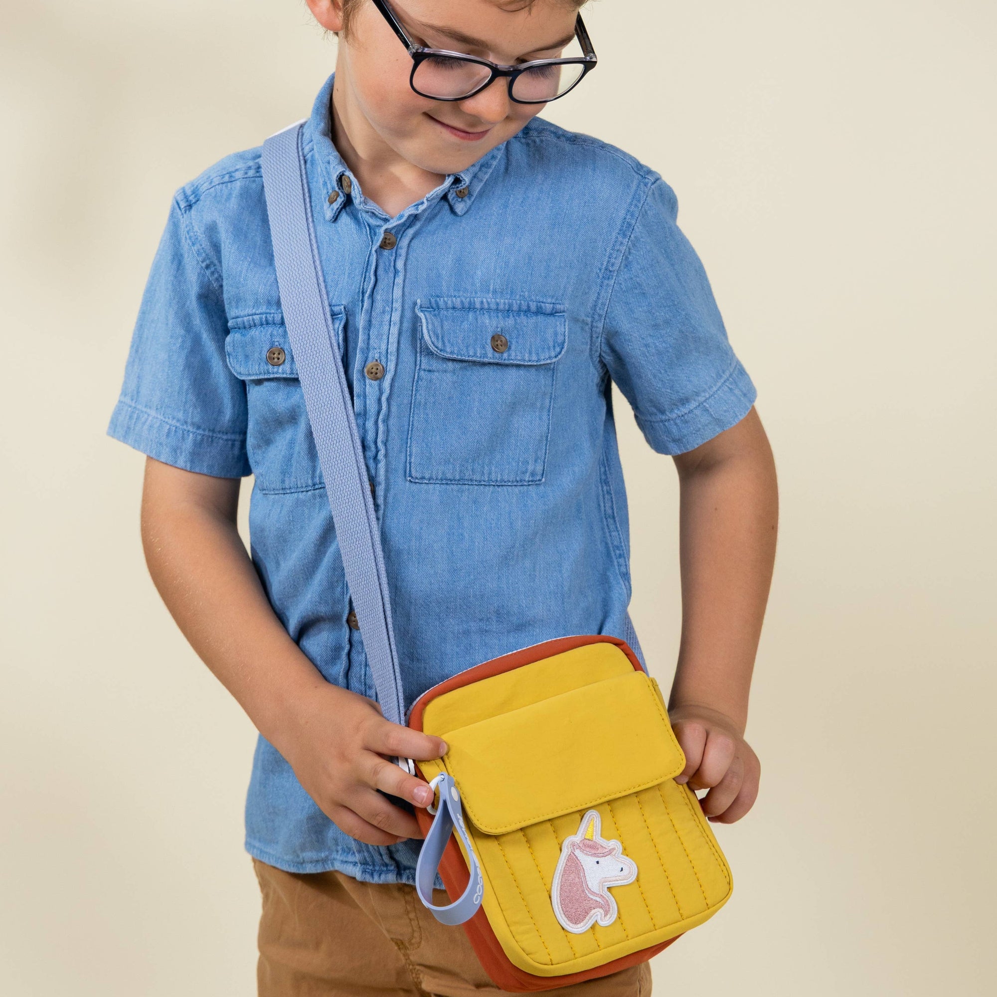 Child wearing glasses and a blue shirt holding a yellow bag with a pink character on a beige background