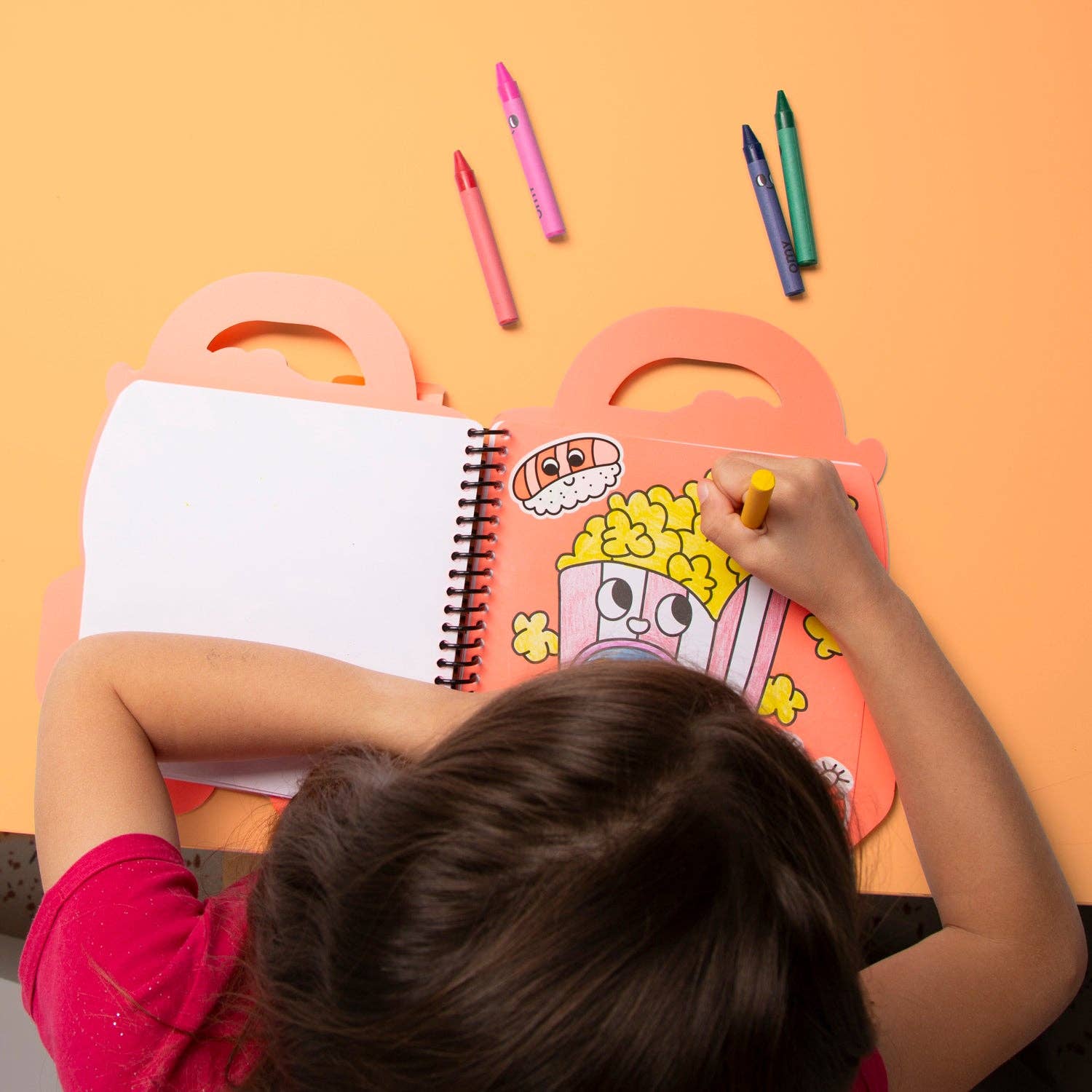 Child coloring in a spiral-bound notebook with colorful crayons on an orange background