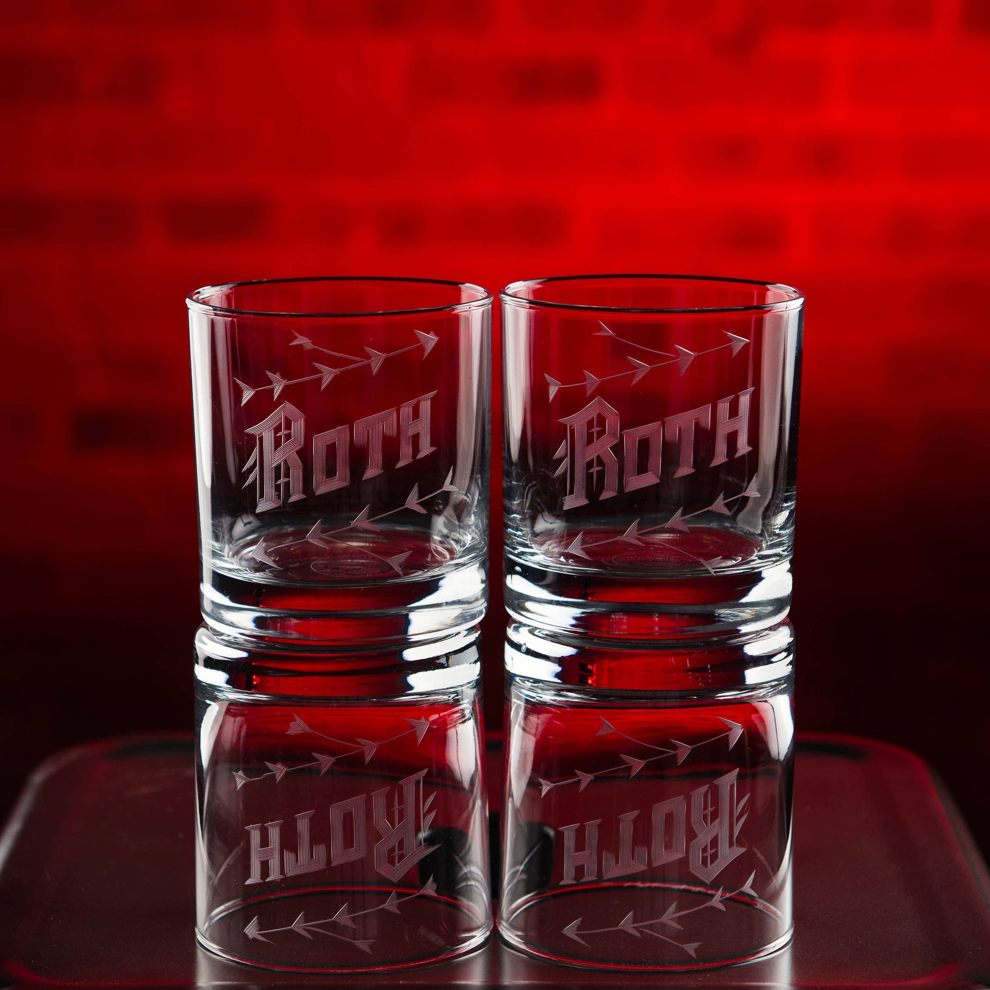 Two clear glasses with 'Roth' engraving on a reflective surface with a red background
